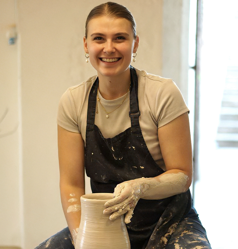 Junge Frau beim Töpfern, lächelnd in einem sommerlichen T-Shirt, an einer Töpferscheibe sitzend und mit Ton auf der Hand, in einem Töpferstudio.