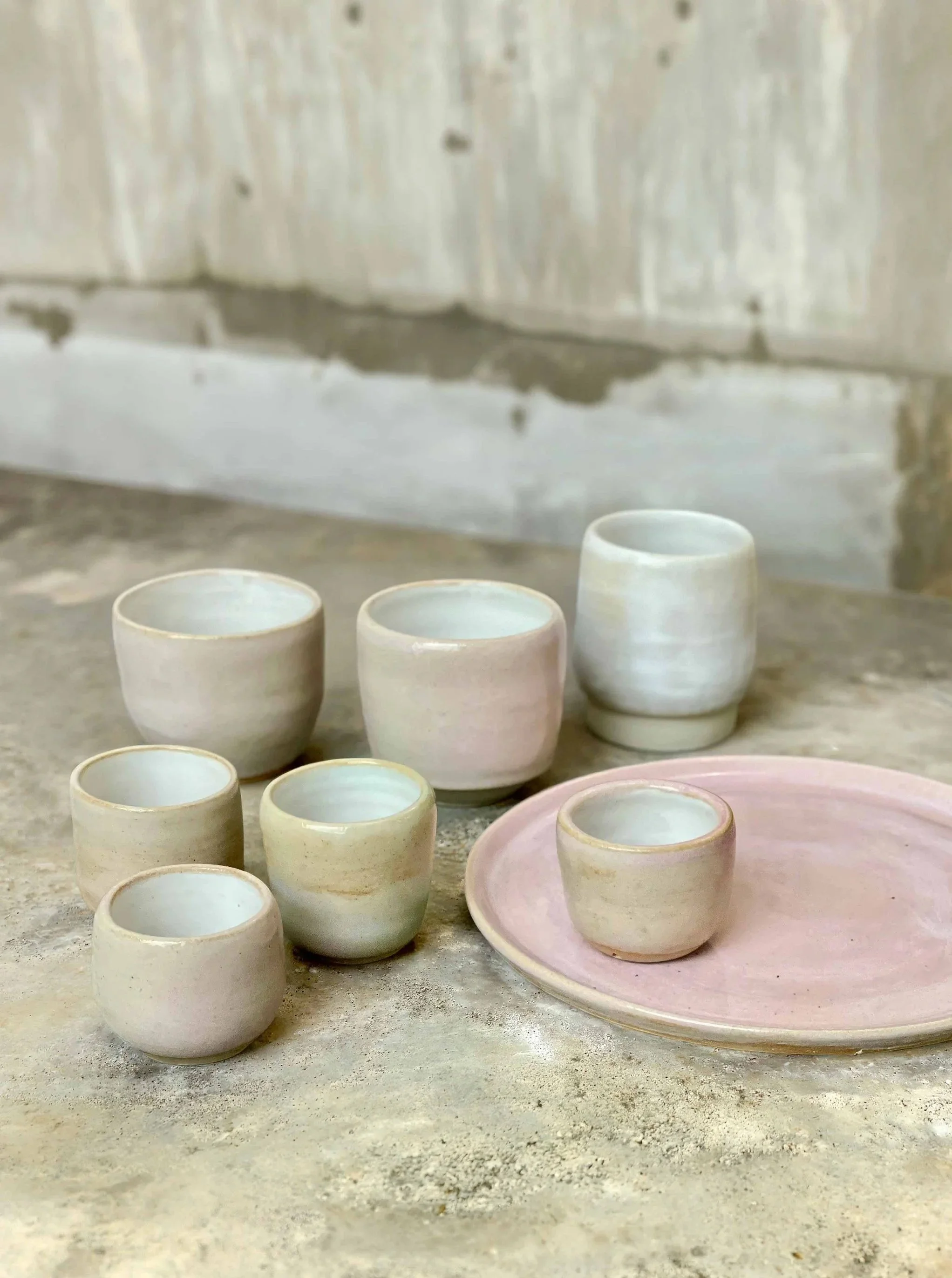 Handgetöpferte rosa-weiße Becher, Tassen, Espresso-Tassen und ein Teller auf einem Betontisch in einem Töpferstudio.