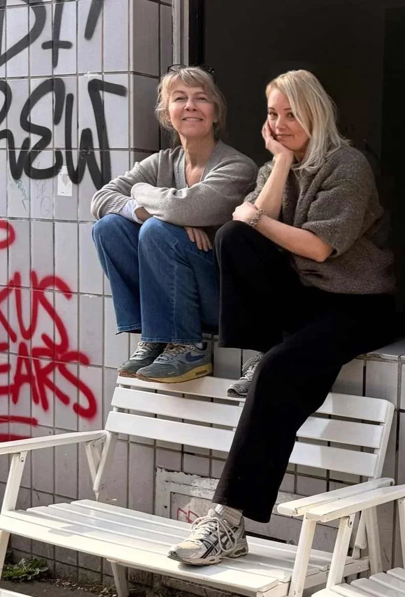 Zwei Frauen sitzen lächelnd im offenen Fenster des Töpferstudios im Erdgeschoss, ihre Füßen sind auf der Lehne einer weißen Holzbank, die vor dem Fenster steht, abgestützt. Die Hauswand zeigt diverse Graffiti.