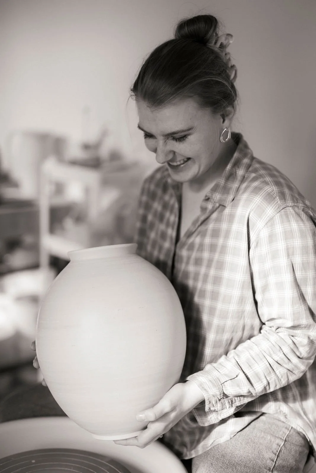 Eine lächelnde Frau sitzt an einer Drehscheibe in einem Töpferstudio und hält eine große, ungebrannte Keramikvase in der Hand.