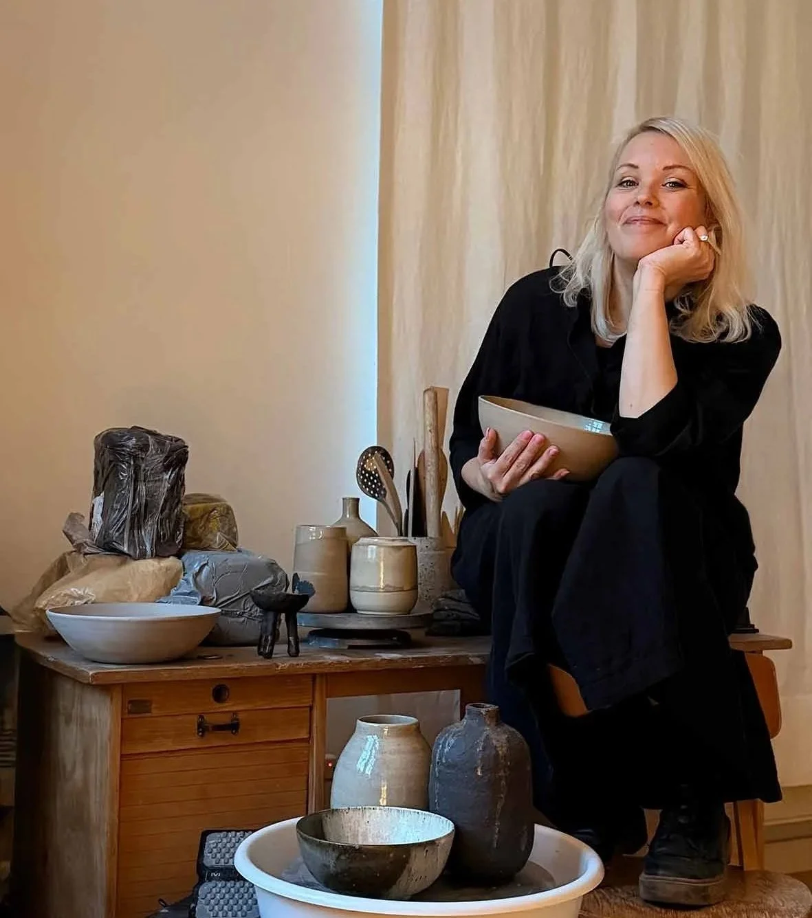 Eine Frau sitzt auf einem Stuhl vor einem Holztisch, auf dem Tonvasen, Schalen und Küchenutensilien aus Keramik stehen. Sie hält eine selbstgetöpferte Schale in der Hand und lächelt in die Kamera.