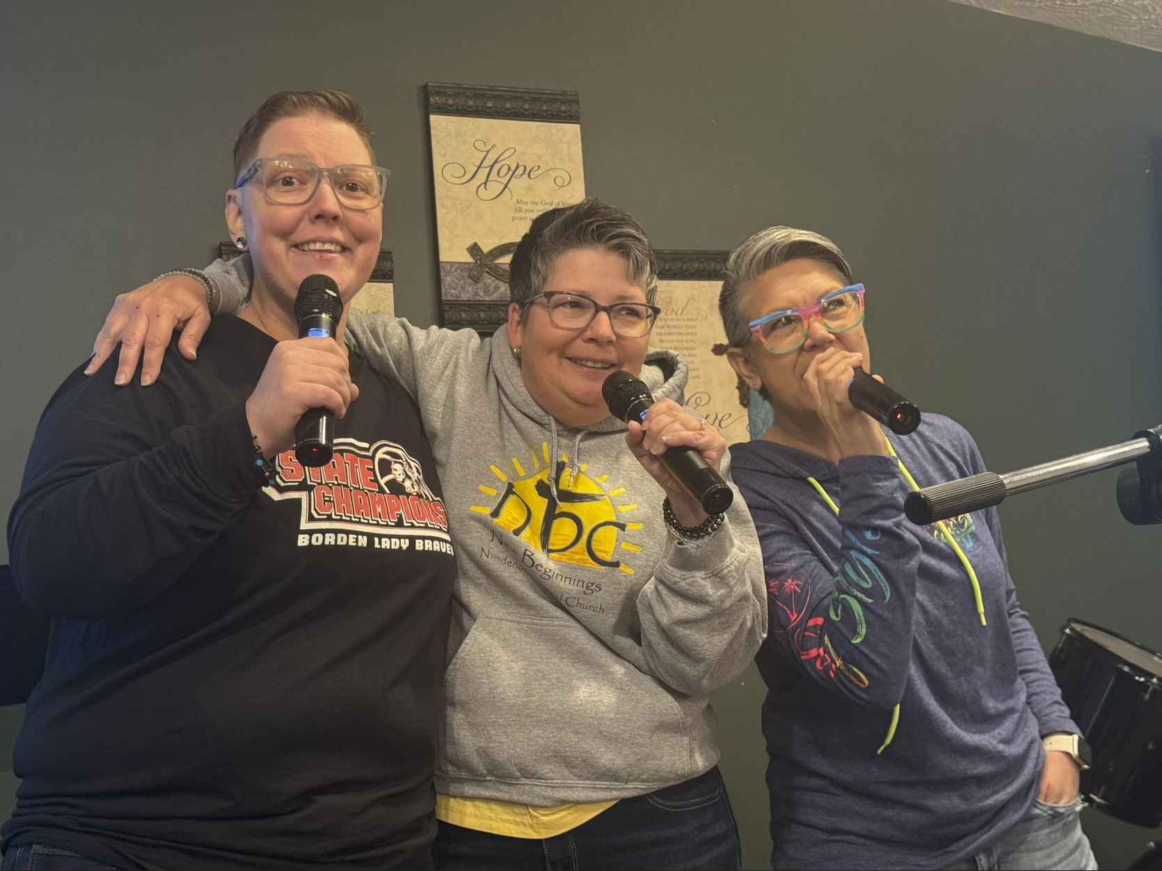 Three women standing close together, holding microphones, with one woman having her arm around the other two, singing or speaking into the microphones. They are in a room with framed signs on the wall behind them.