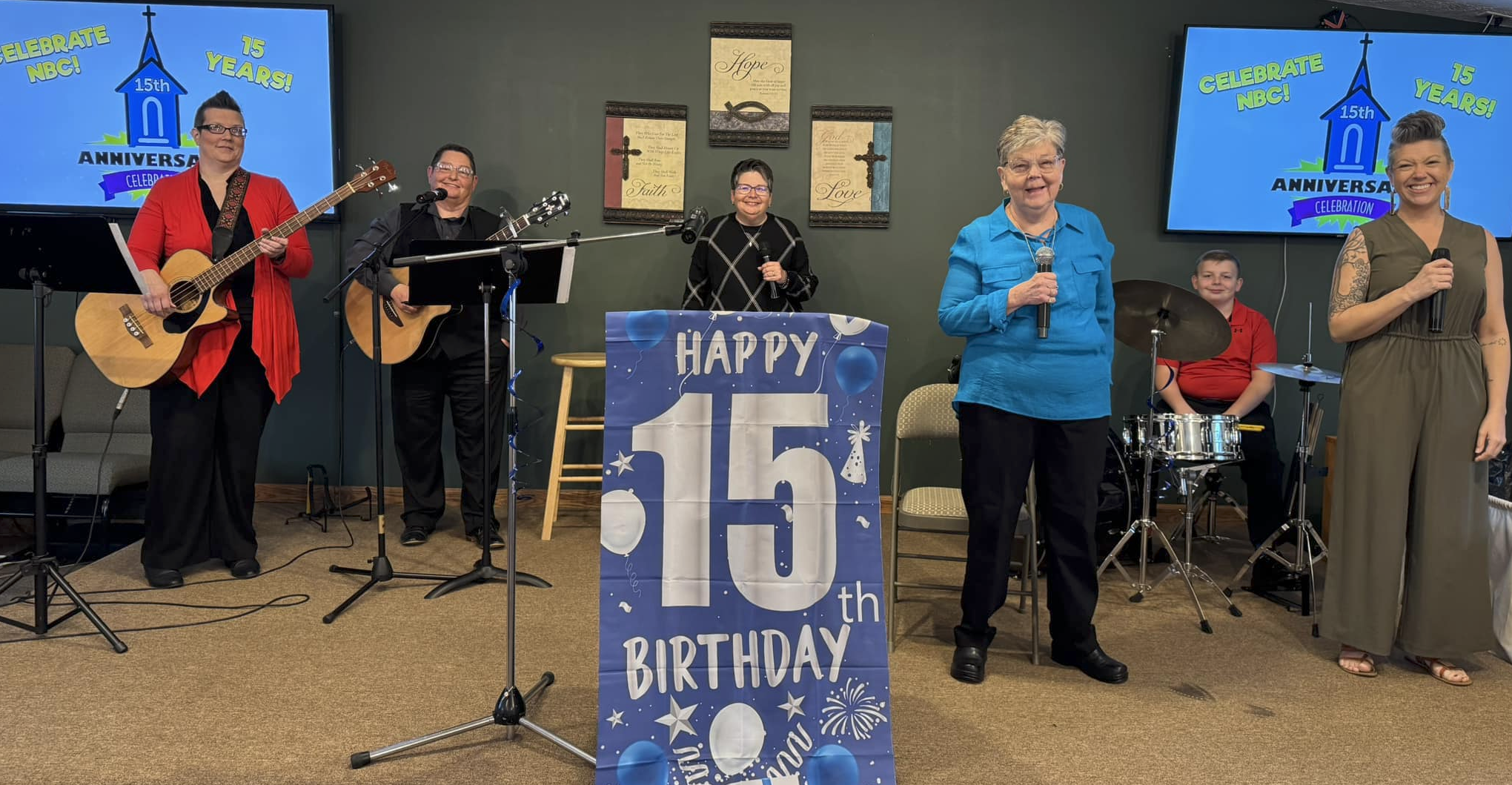 Group of seven people celebrating a 15th birthday with a musical performance and a large birthday poster. There are two women playing guitars, a woman singing, a woman holding a microphone, a young boy playing drums, and two women speaking into microphones. Two TV screens display a graphic celebrating NBC's 15th anniversary.