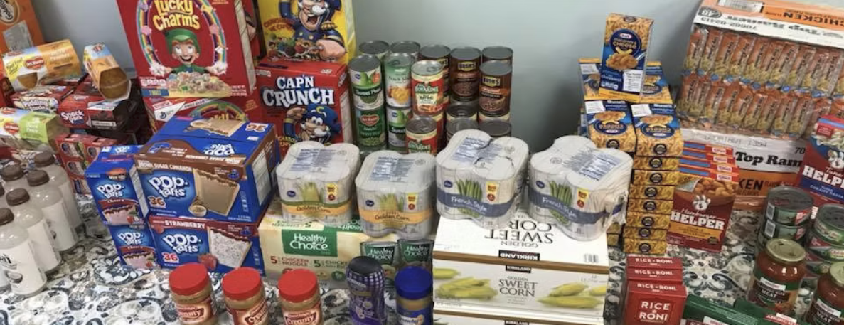 Assorted groceries on a table including cereal boxes, canned vegetables, snack packs, bottles of vitamins, and spice jars.