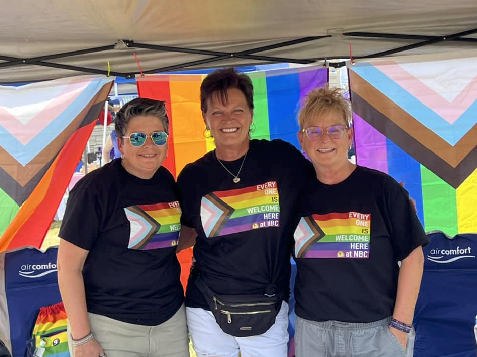Three women smiling and wearing black T-shirts with rainbow-themed welcome signs, standing in front of rainbow flags and decorations at an outdoor pride event.