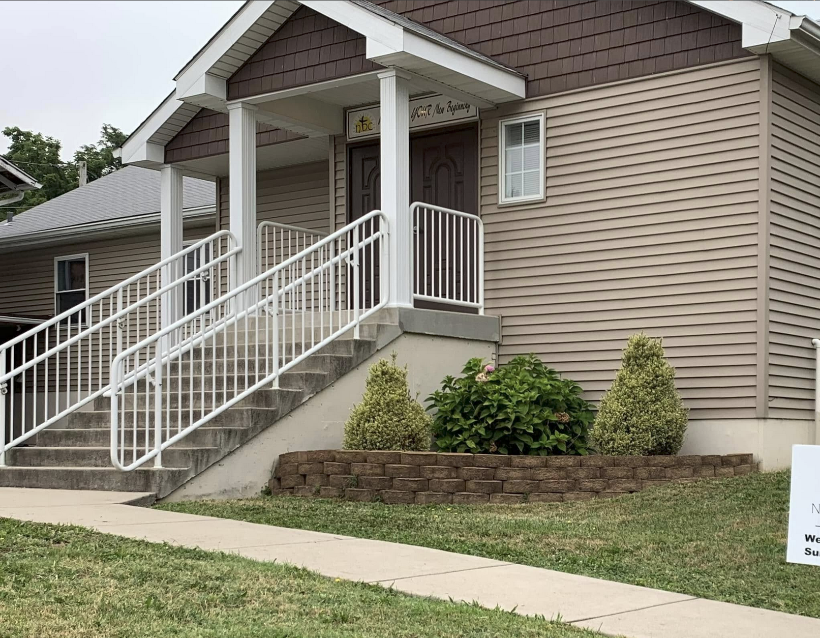 Front view of a beige house with a small porch, white railing, and stairs leading up to the entrance. There are two shrubbery plants and a hydrangea flower in a brick-bordered garden bed near the stairs.