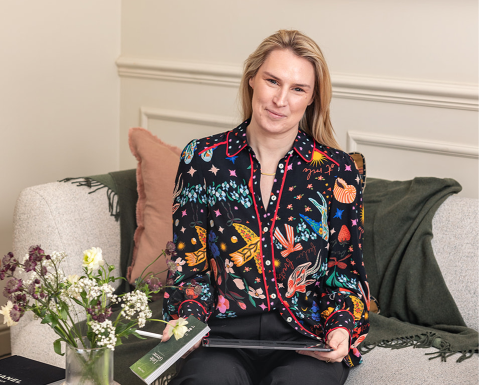 Rosa Wynn Jones sits on a beige sofa with pink and green throw pillows, holding a laptop, next to a glass vase of mixed flowers, in a room with cream walls and white trim, wearing a colourful patterned blouse.
