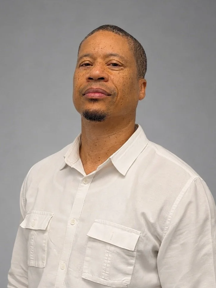 A man with short hair and a goatee, wearing a white collared shirt, standing against a gray background.