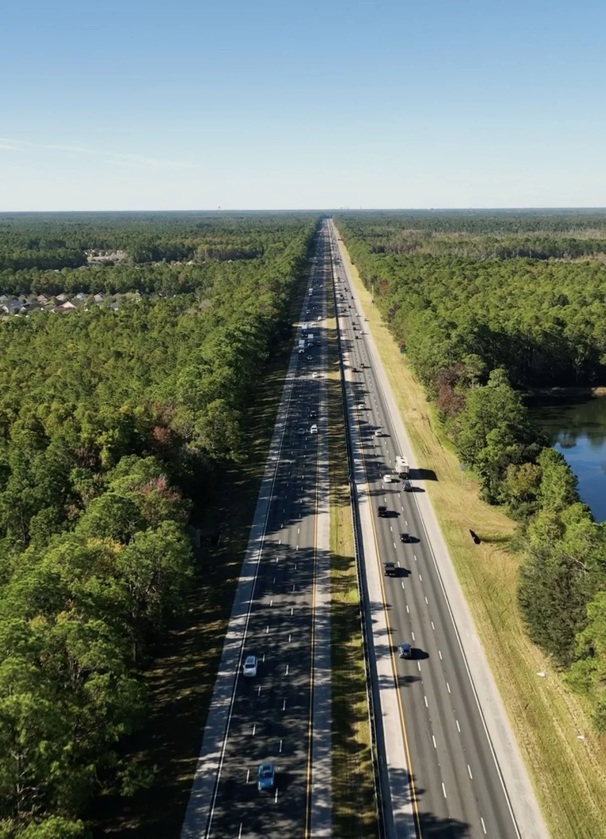 An aerial view of a divided highway with cars traveling in both directions, surrounded by dense green trees and a small body of water on the right side under a clear blue sky.