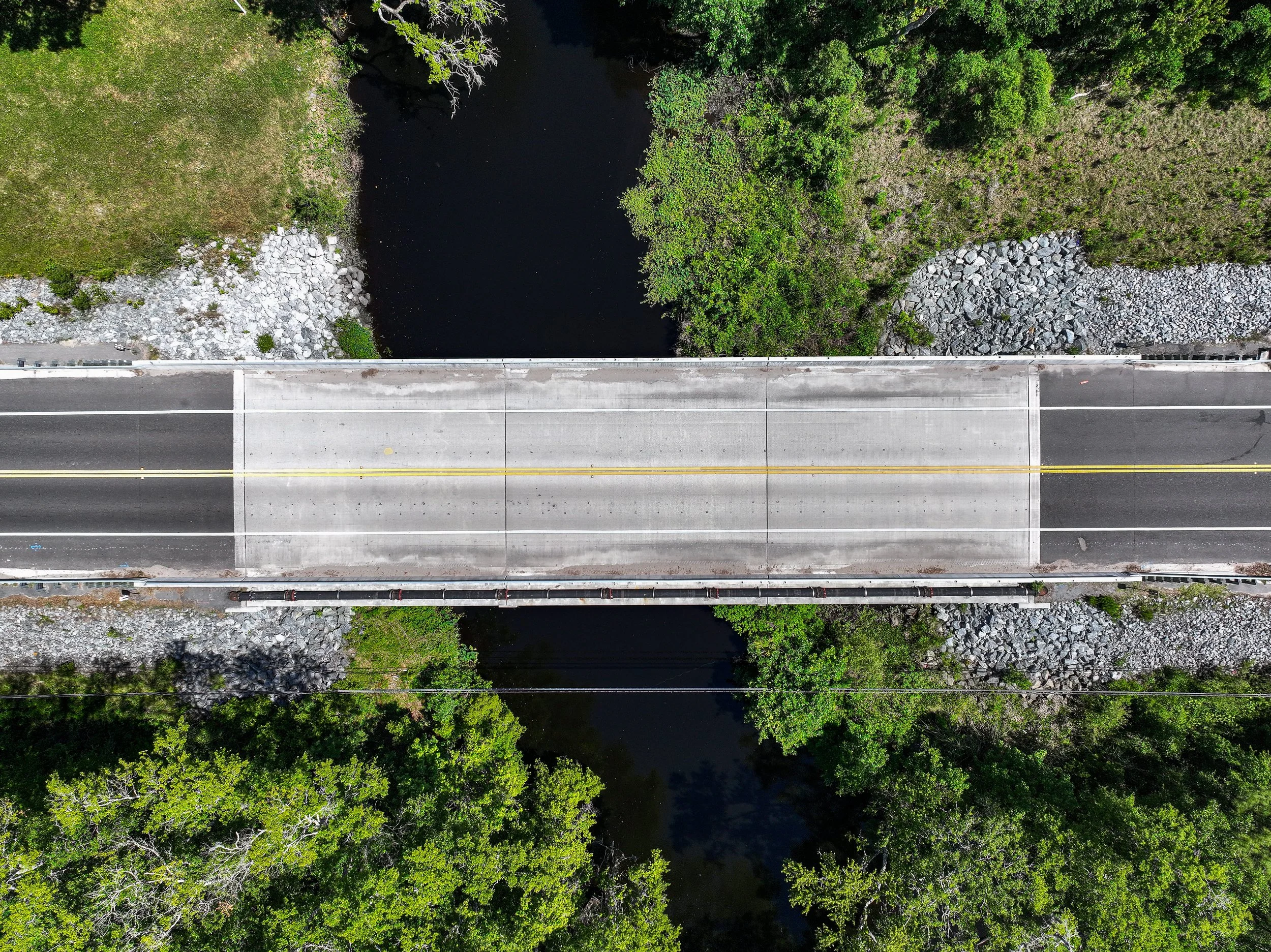An overhead aerial view of a bridge crossing over a waterway with lush green trees on each side.