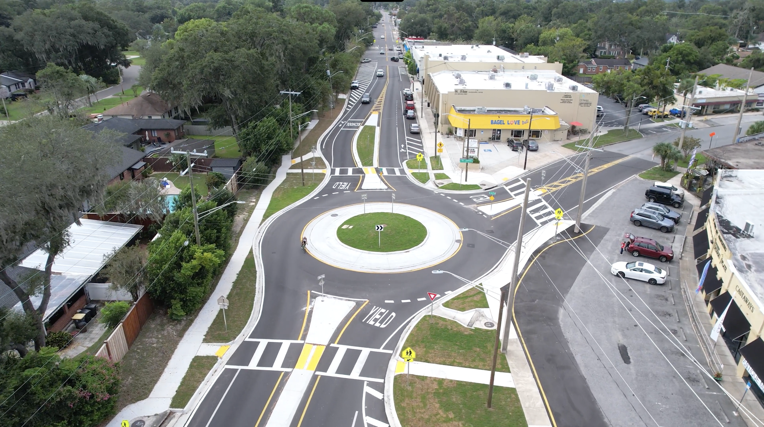 Aerial view of a new traffic roundabout with a circular island and bike lane, surrounded by roads, cars, and green trees, in a suburban area with shops and residential homes.
