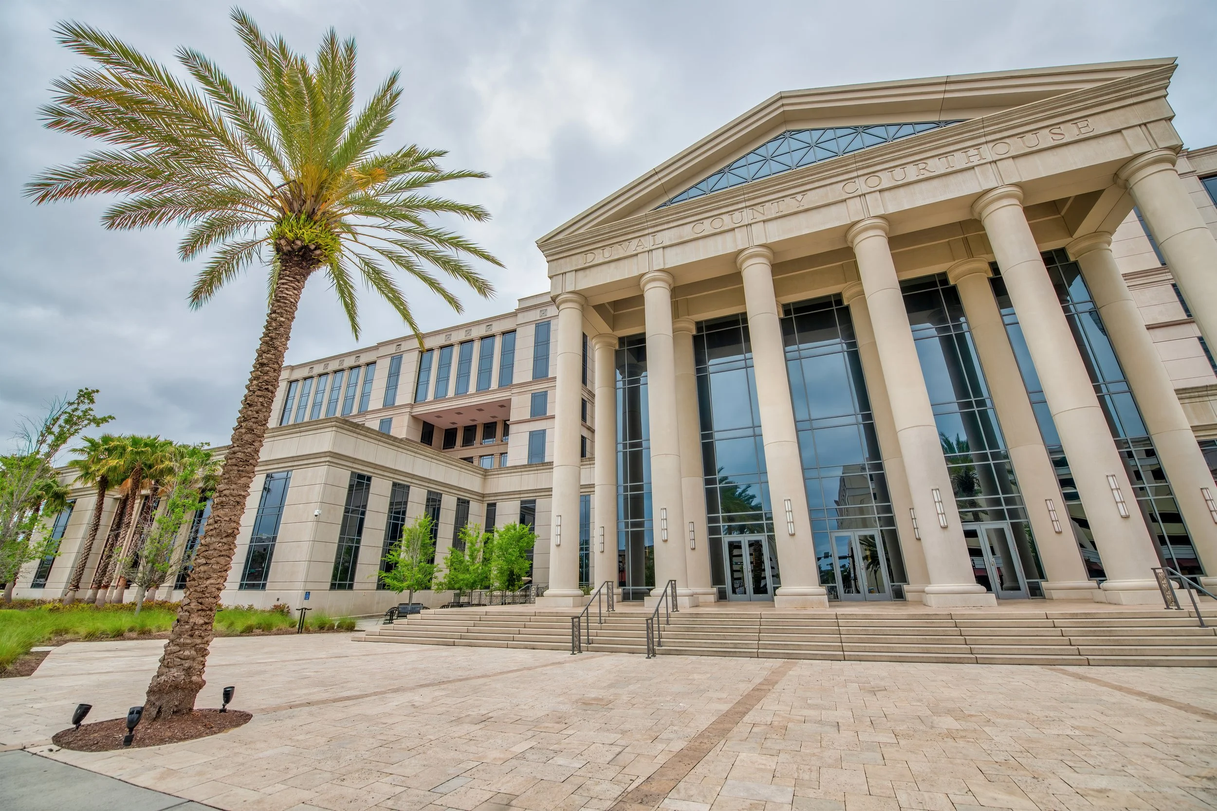 Front exterior view of Duval County Courthouse with tall pillars, glass windows, steps, and a palm tree in the foreground.