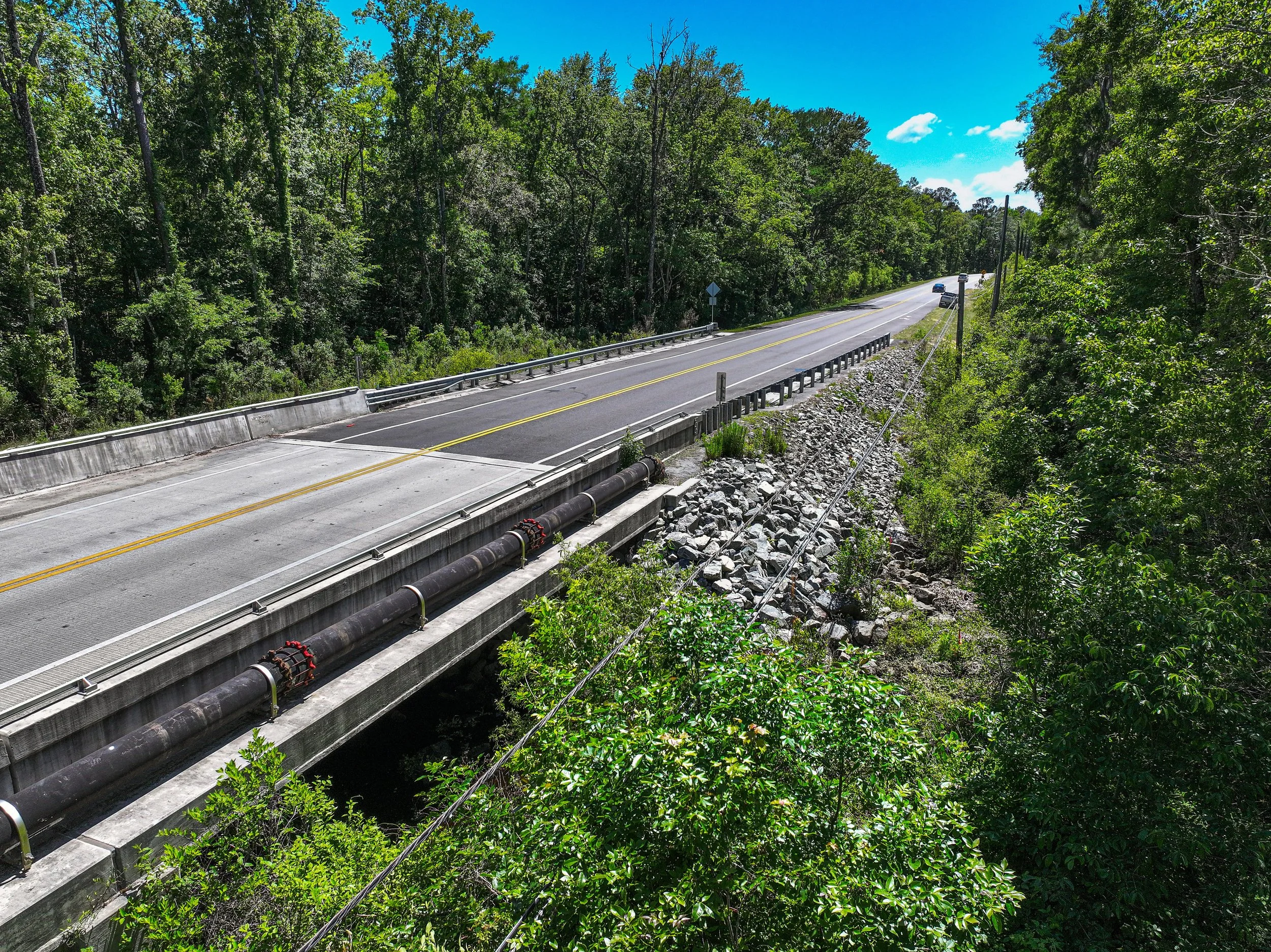 A two-lane road with a double yellow line in the center, cutting through a dense green forest on both sides, with a blue sky and a few clouds overhead.