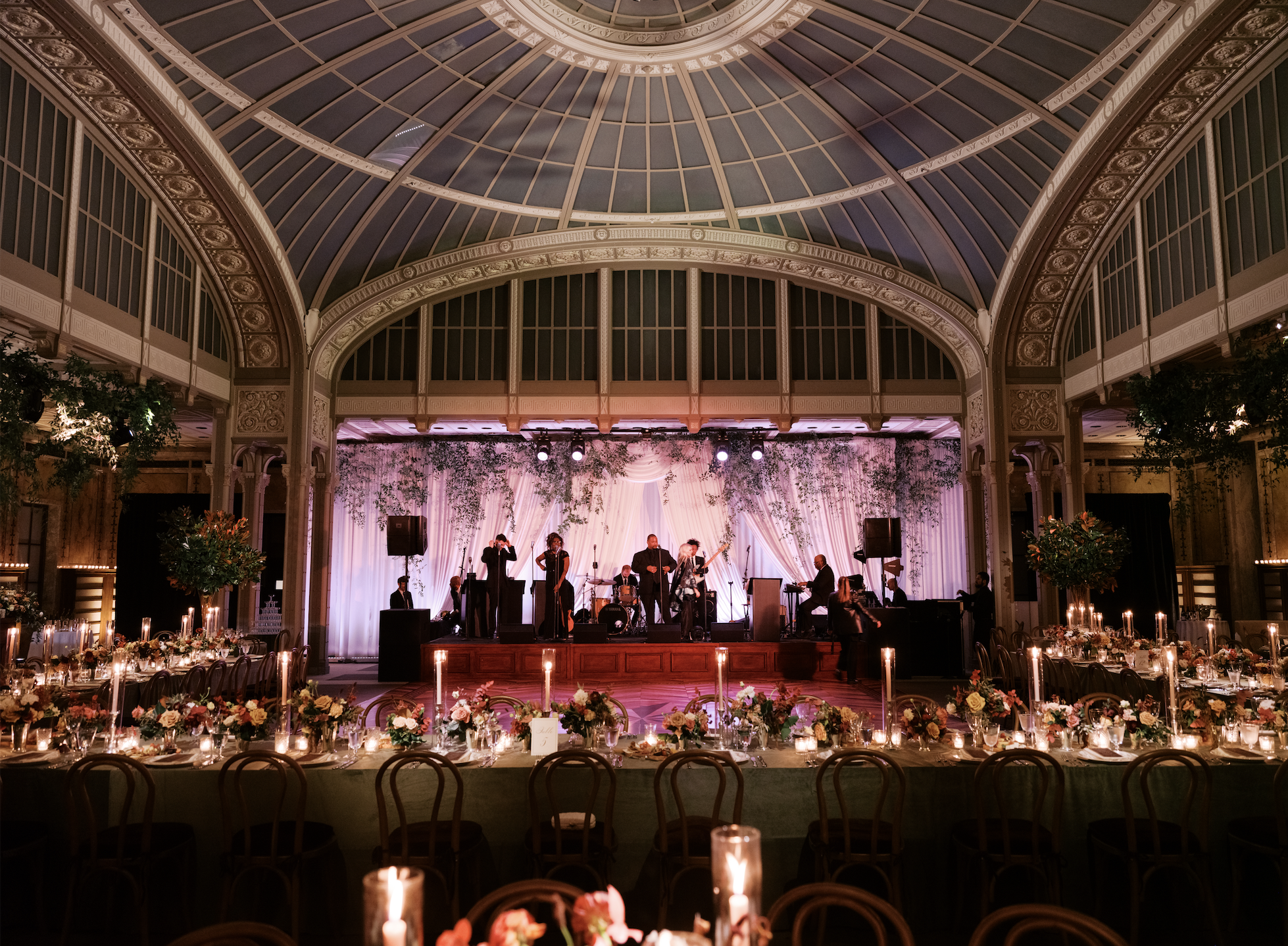 A stage with a live band performing in a decorated banquet hall. The hall features a domed ceiling, lace curtains, and floral arrangements, with tables set for dining and candles lighting the foreground.