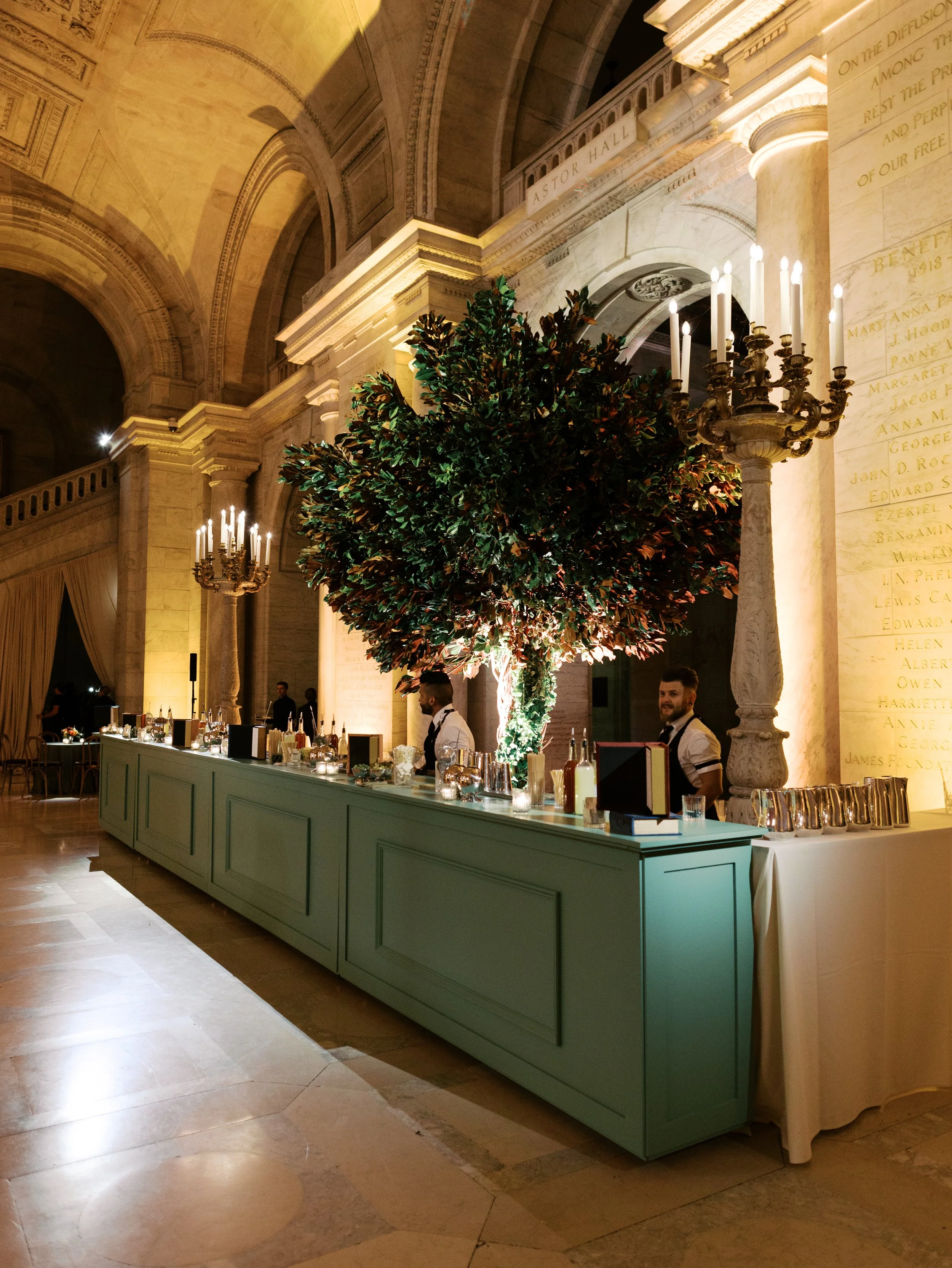 Elegant reception bar with a large illuminated tree centerpiece, inside a historic building with high vaulted ceilings and classical architectural details.