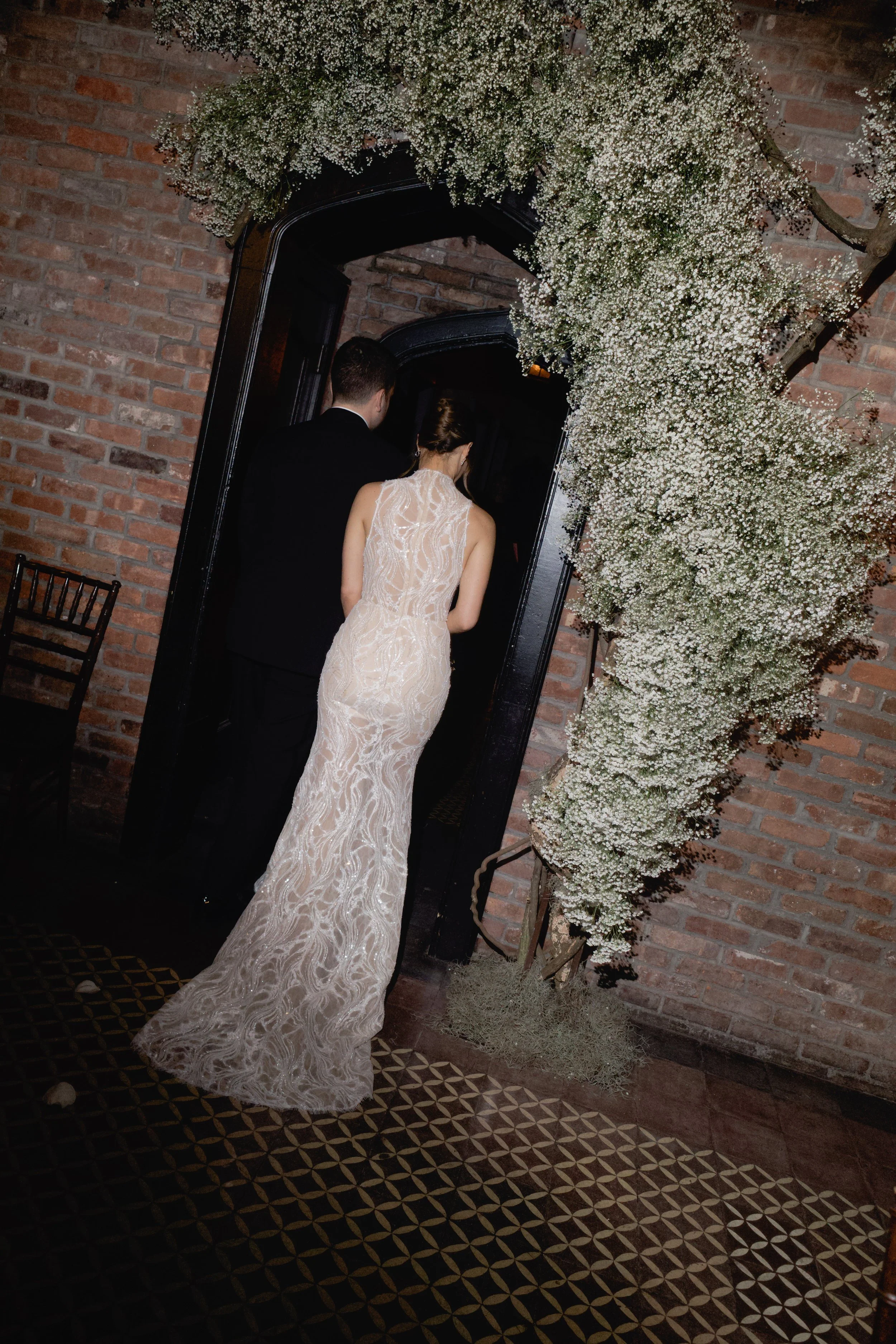 A bride in a white lace wedding gown and a groom in a black suit enter a room through a black door framed with white baby's breath flowers and greenery, with a brick wall in the background.