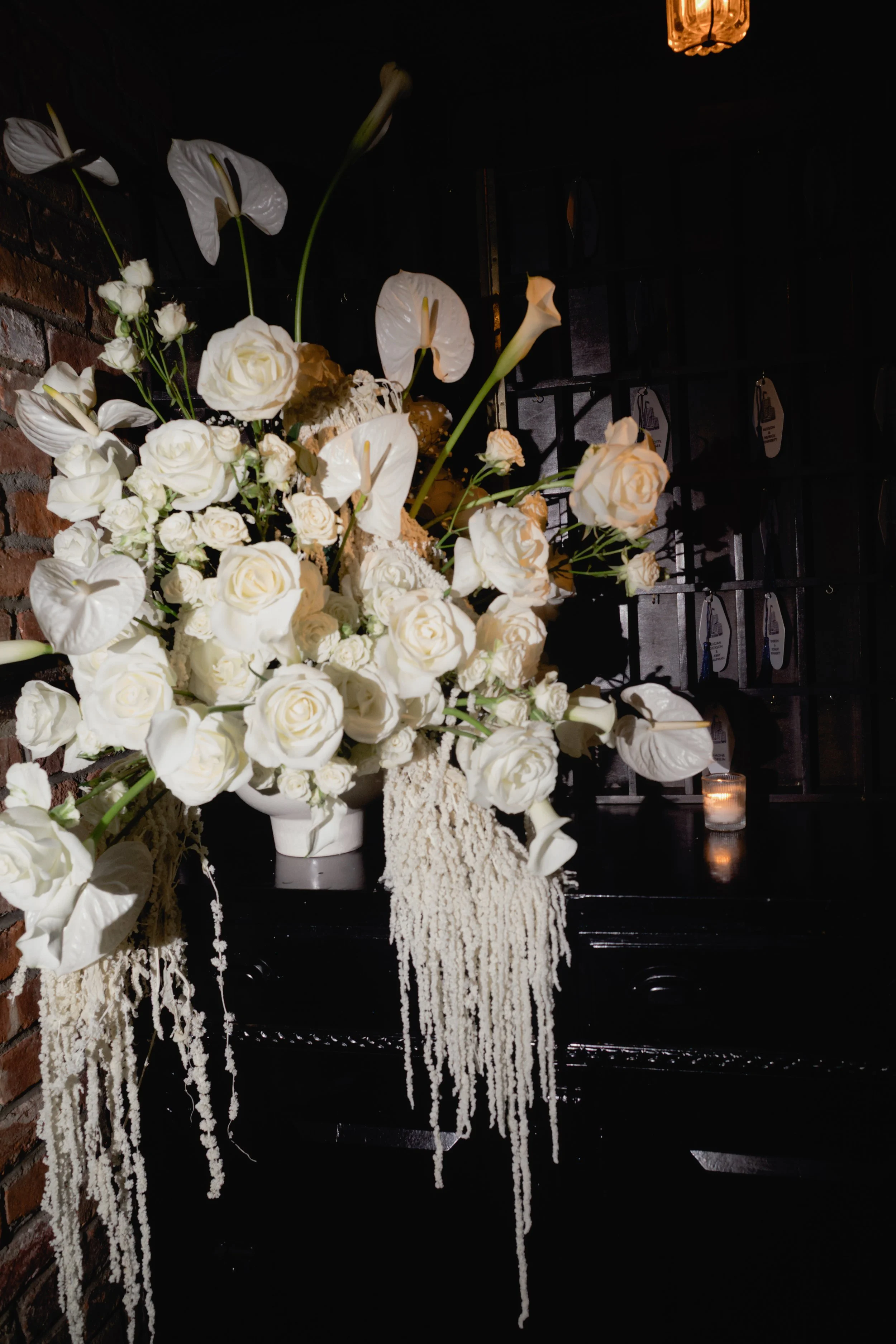 Elegant white floral arrangement with roses, calla lilies, and hanging amaranth flowers on a dark background, illuminated by candlelight.