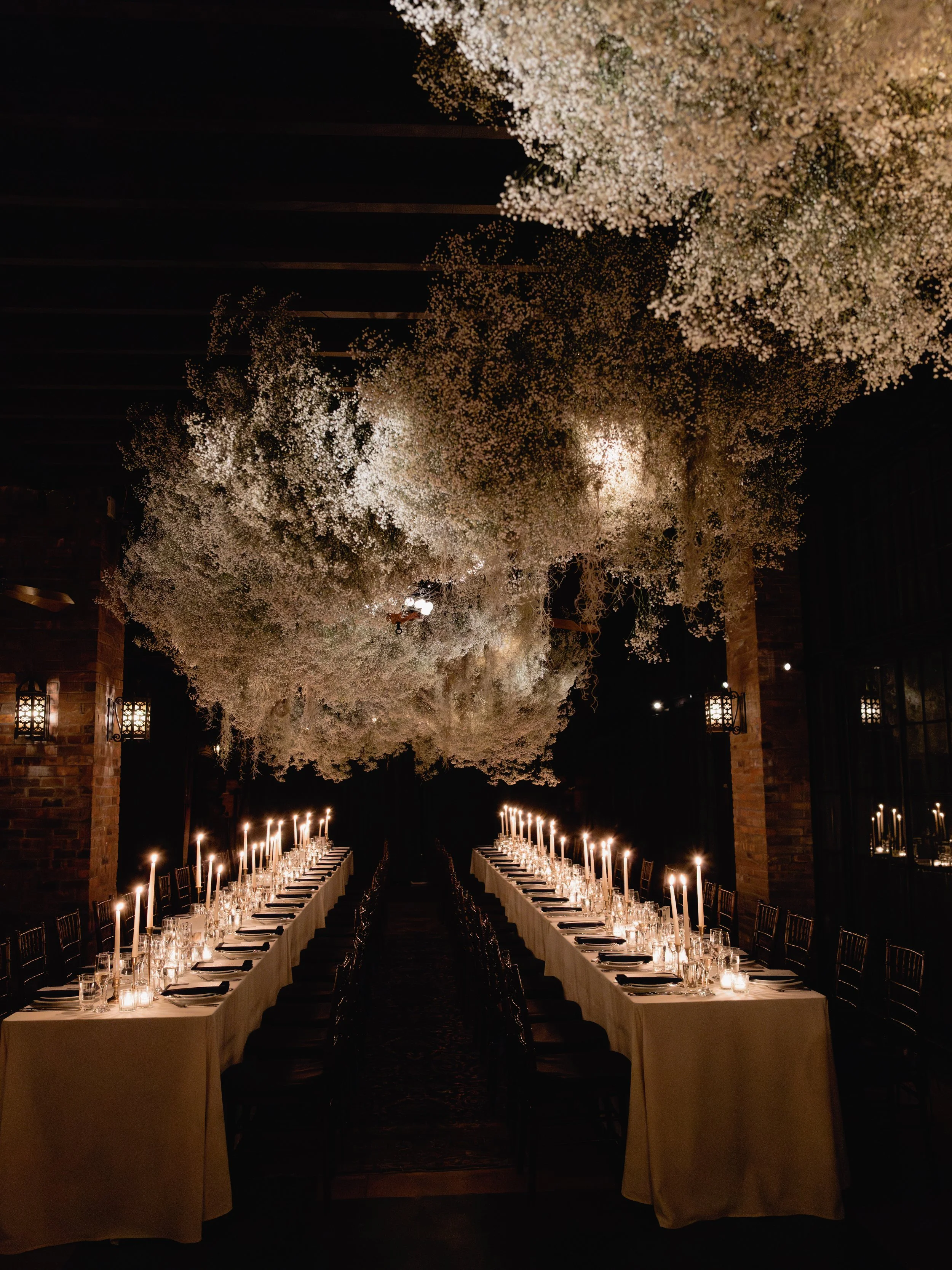 A dimly lit banquet hall decorated with hanging white floral arrangements and long tables set for a dinner with candles and glassware.