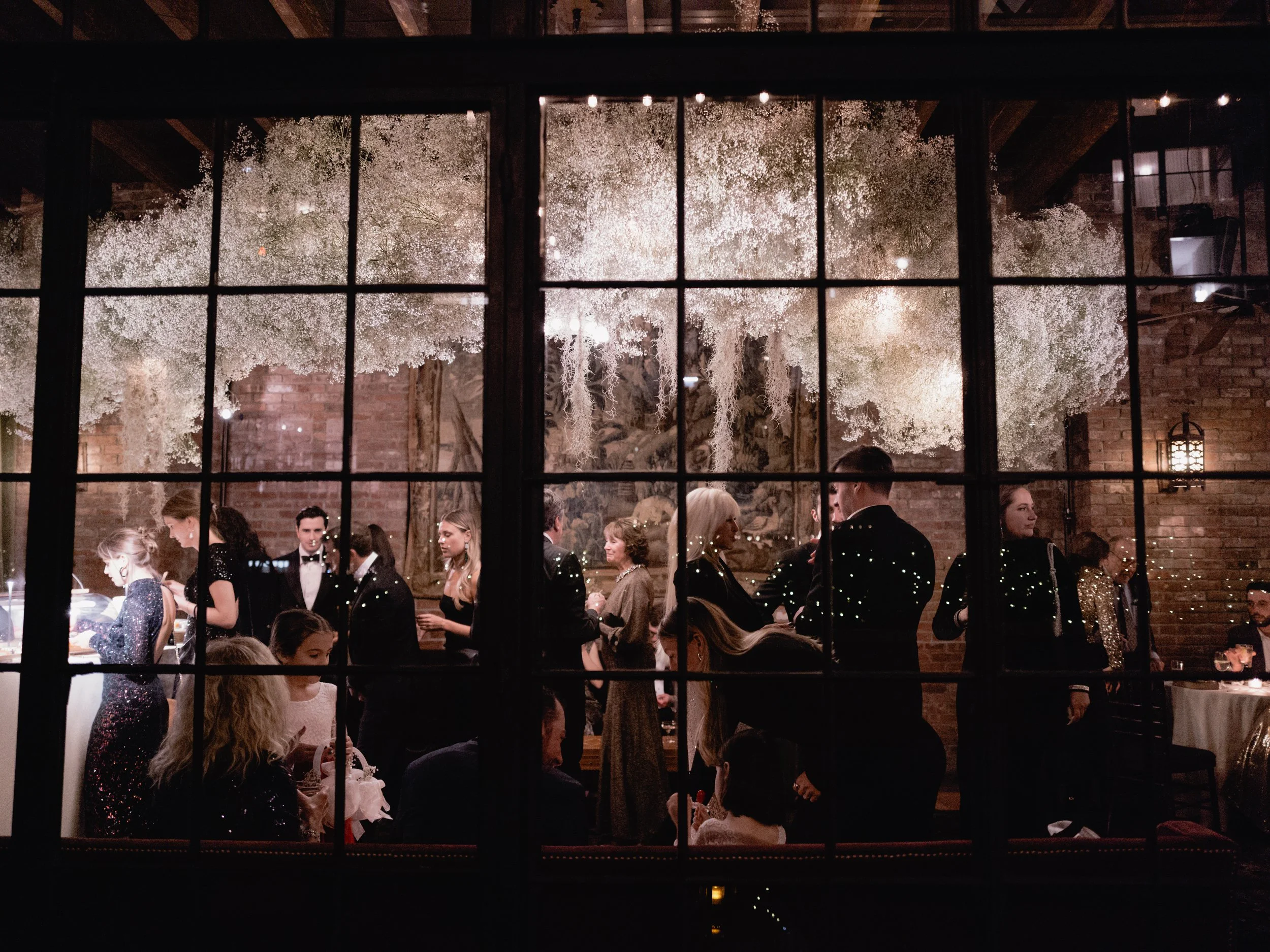 people gathered in a dimly lit room with exposed brick walls, decorated with white floral arrangements and hanging lights, seen through a window with grid panes