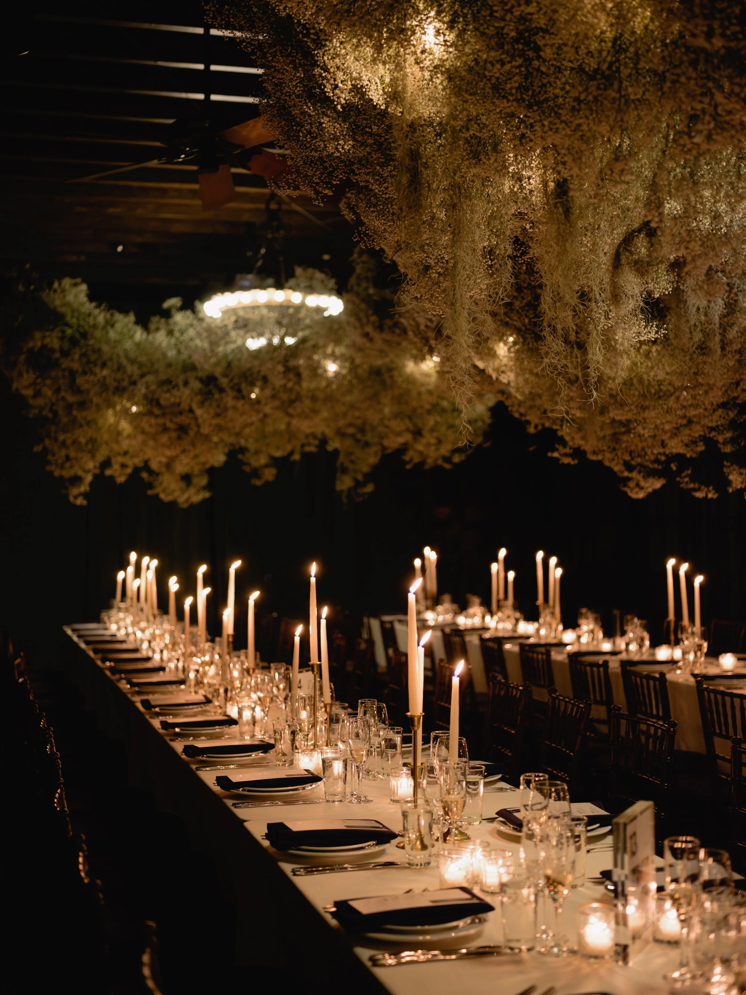 A long banquet table decorated with tall white candles, candles in glass holders, and preset dinnerware, with floral arrangements hanging upside down above, in a dimly lit setting.
