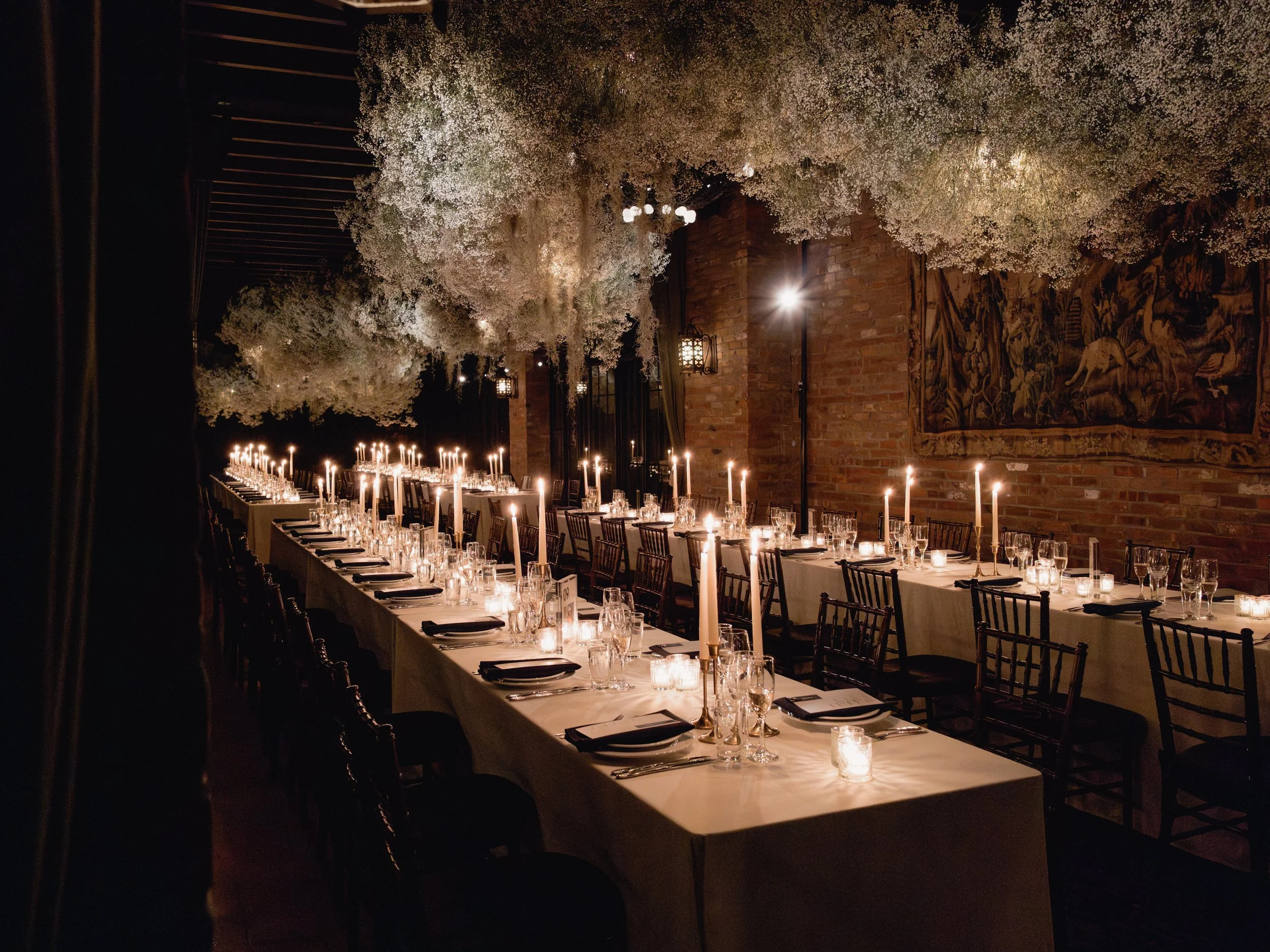 Long banquet table in a dimly lit room with white floral arrangements overhead, lit candles, and dinnerware set for a formal event.