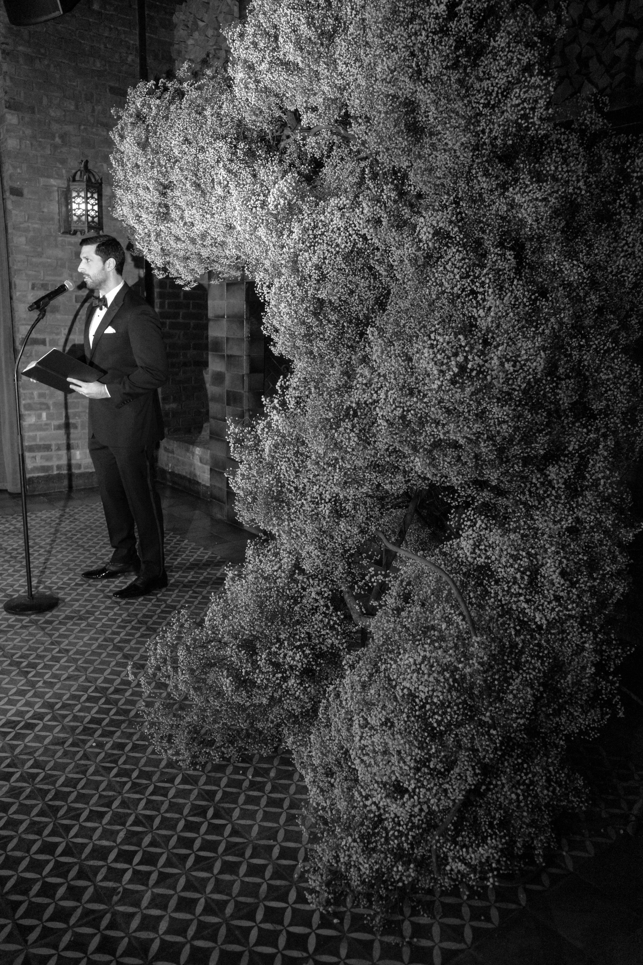 A man in a tuxedo speaking into a microphone at a formal indoor event, with a large floral arrangement of baby's breath and a wall-mounted lantern in the background.