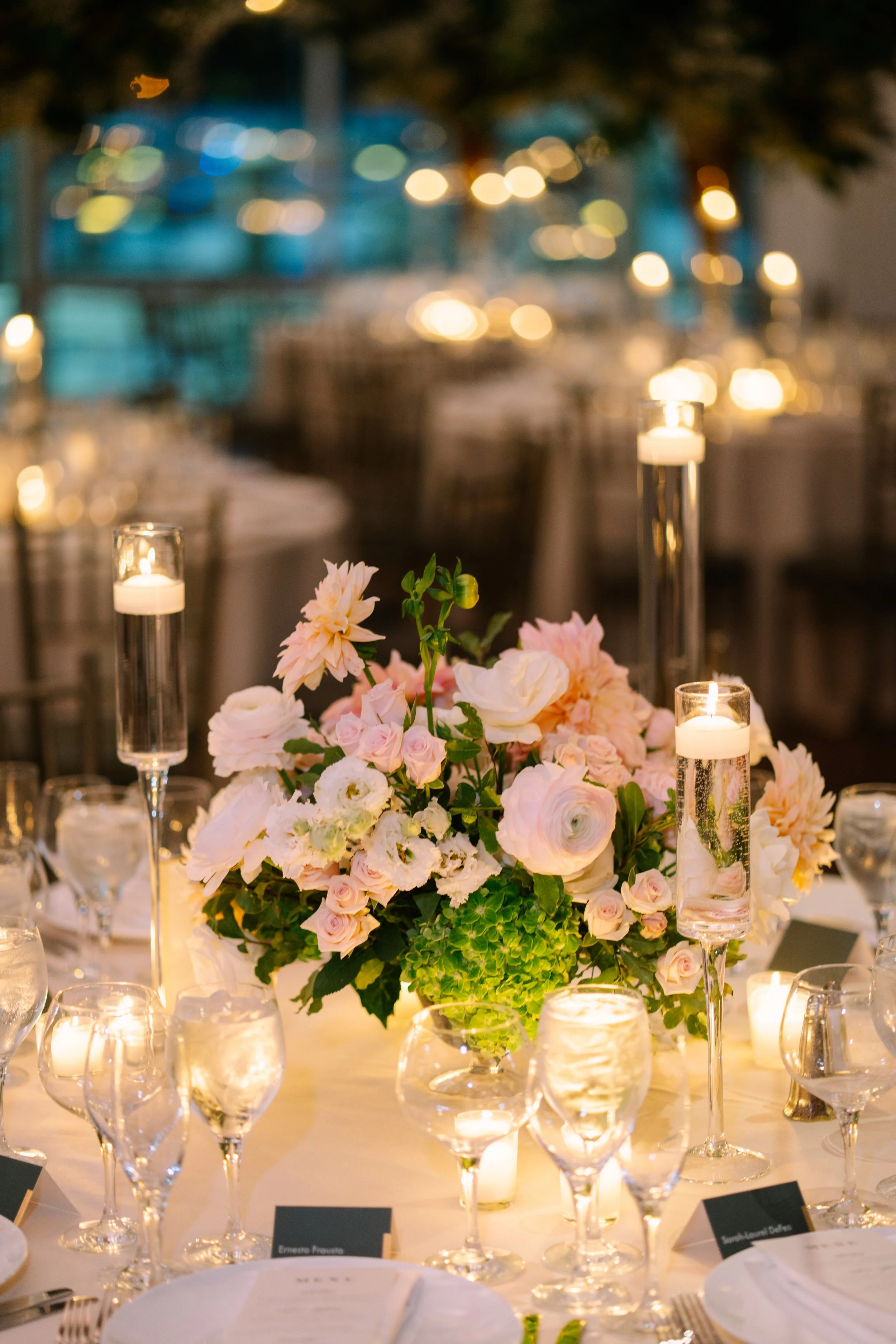 A wedding or event table setup with a floral centerpiece featuring pink and white flowers, surrounded by multiple candle holders with lit candles and glassware.