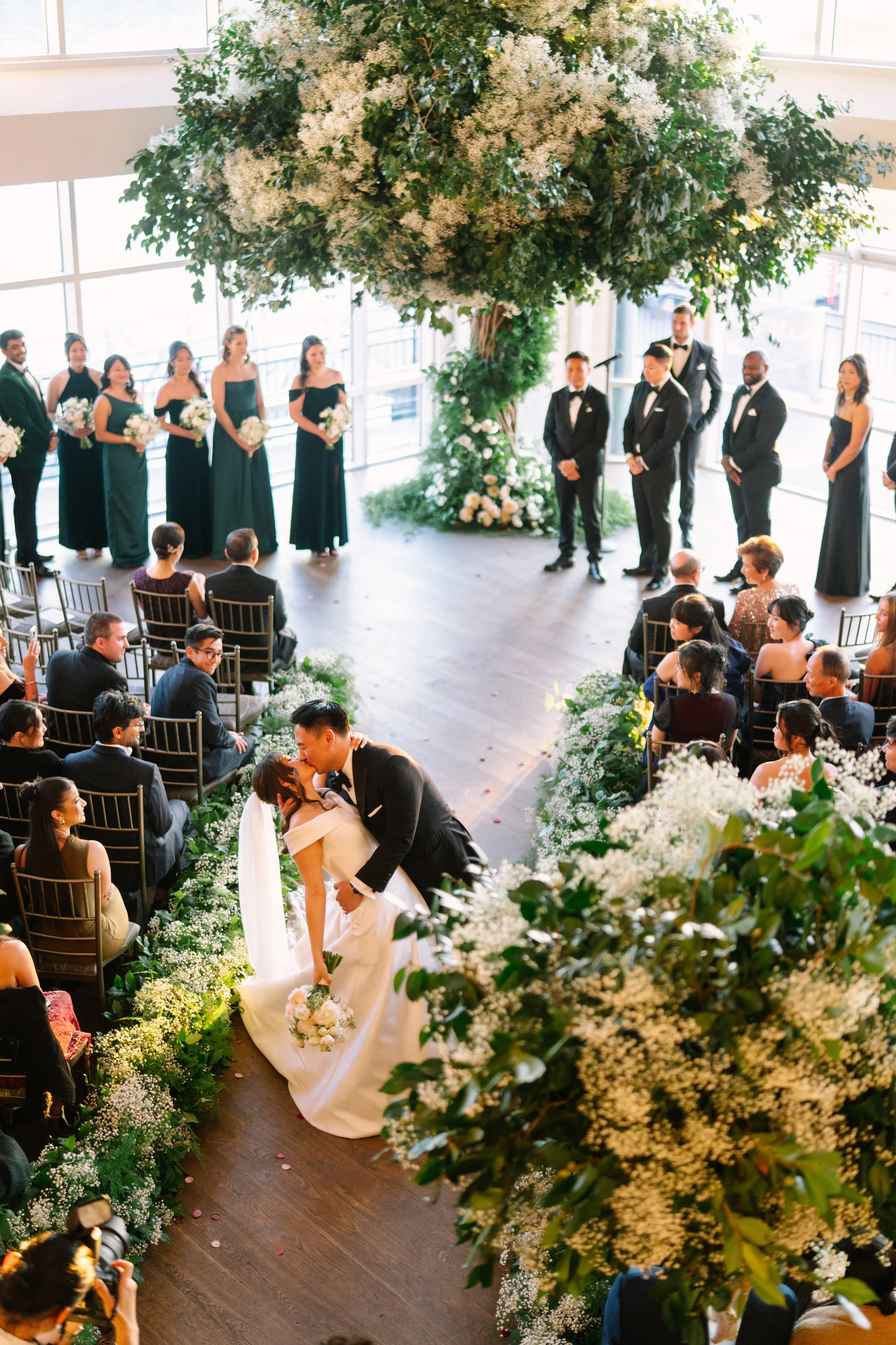A wedding ceremony in a bright, modern indoor space with large windows, featuring a couple kissing at the altar surrounded by flowers, with the wedding party standing behind them and guests seated on either side.