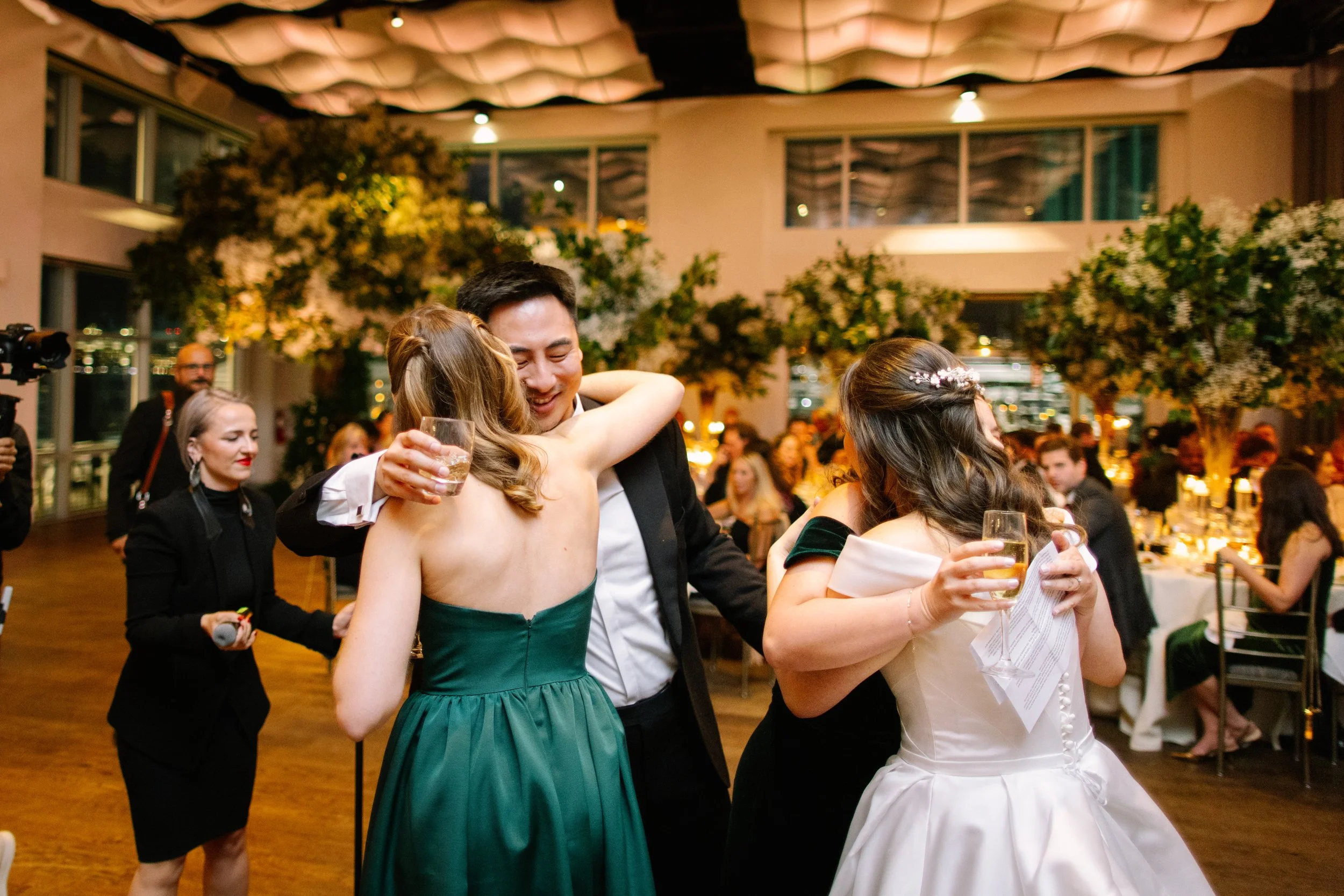 People hugging and celebrating at a wedding reception, with tables and floral decorations in the background.