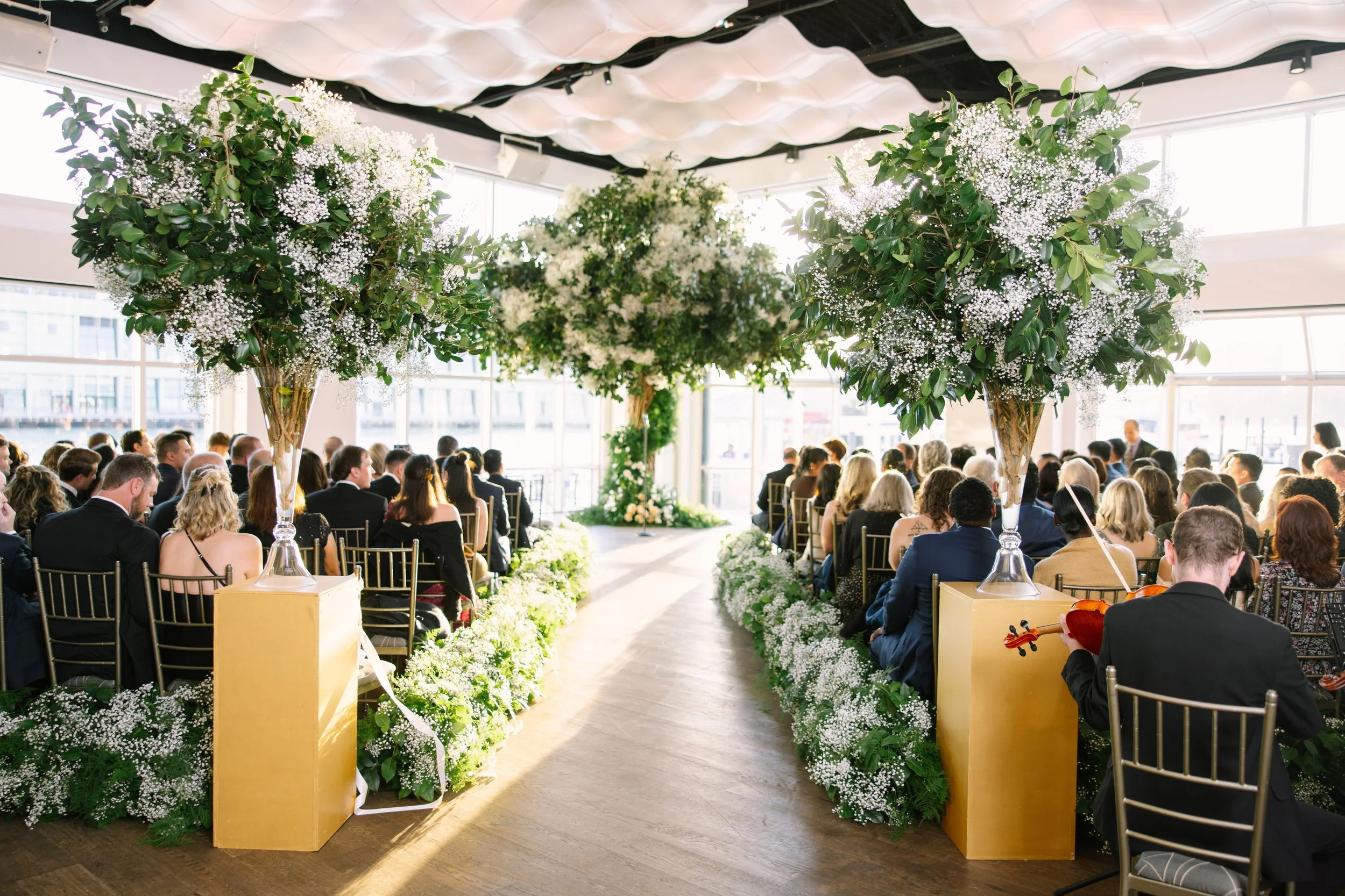 Elegant indoor wedding ceremony with rows of guests seated on either side of a central aisle, decorated with large floral arrangements of greenery and white flowers, under a decorative ceiling, with natural light coming through large windows.