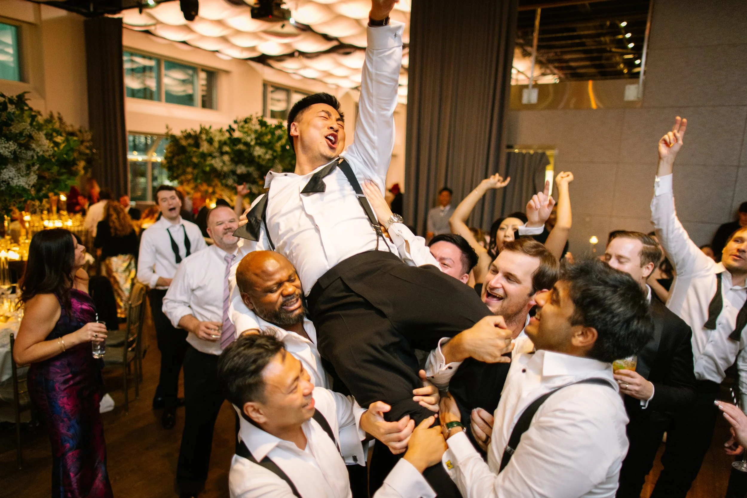 Group of people celebrating, lifting a man in a tuxedo in the air at a party or wedding reception