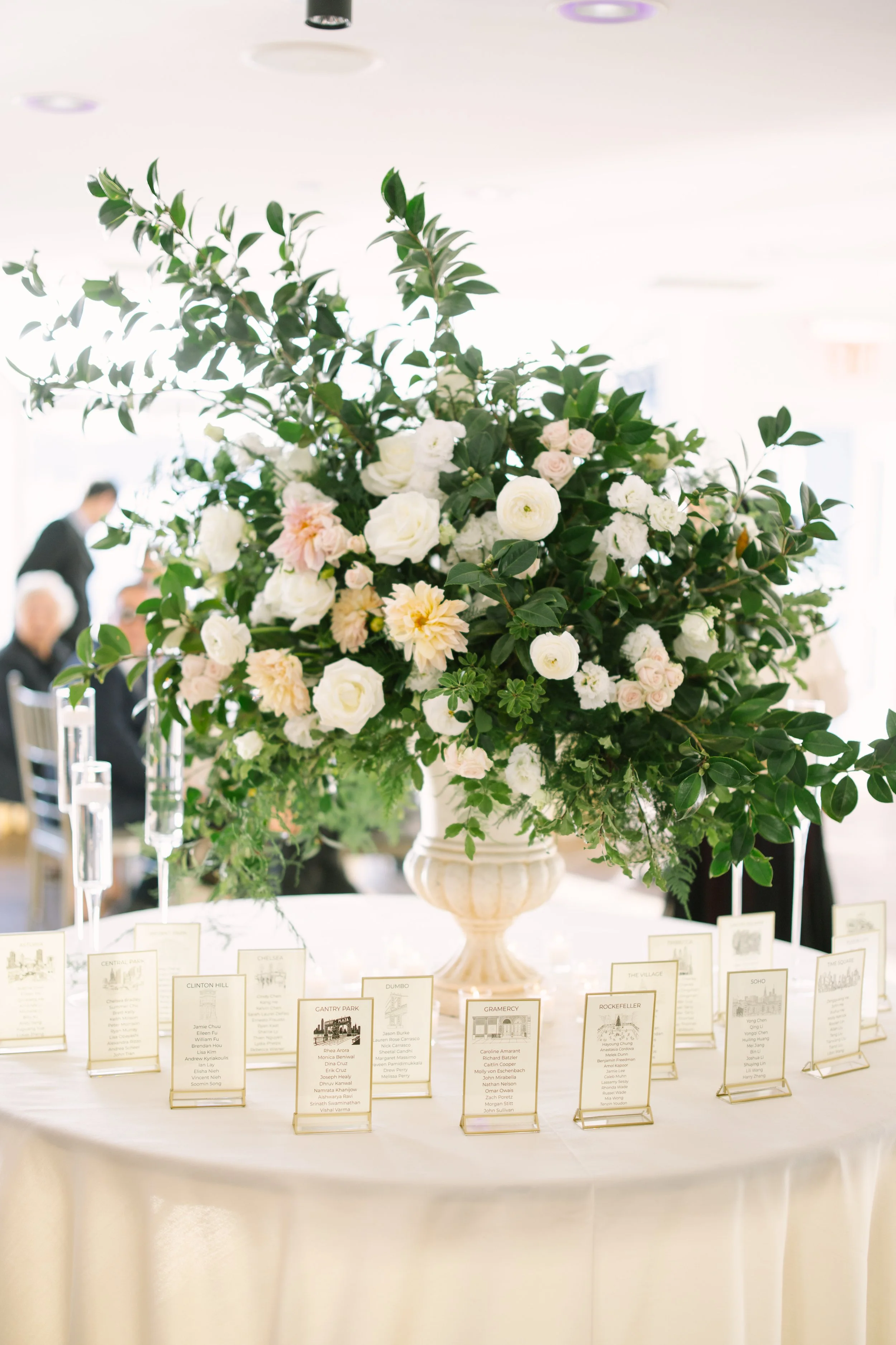 Large centerpiece with white and light pink flowers on a table at an event, with small seating cards in front.