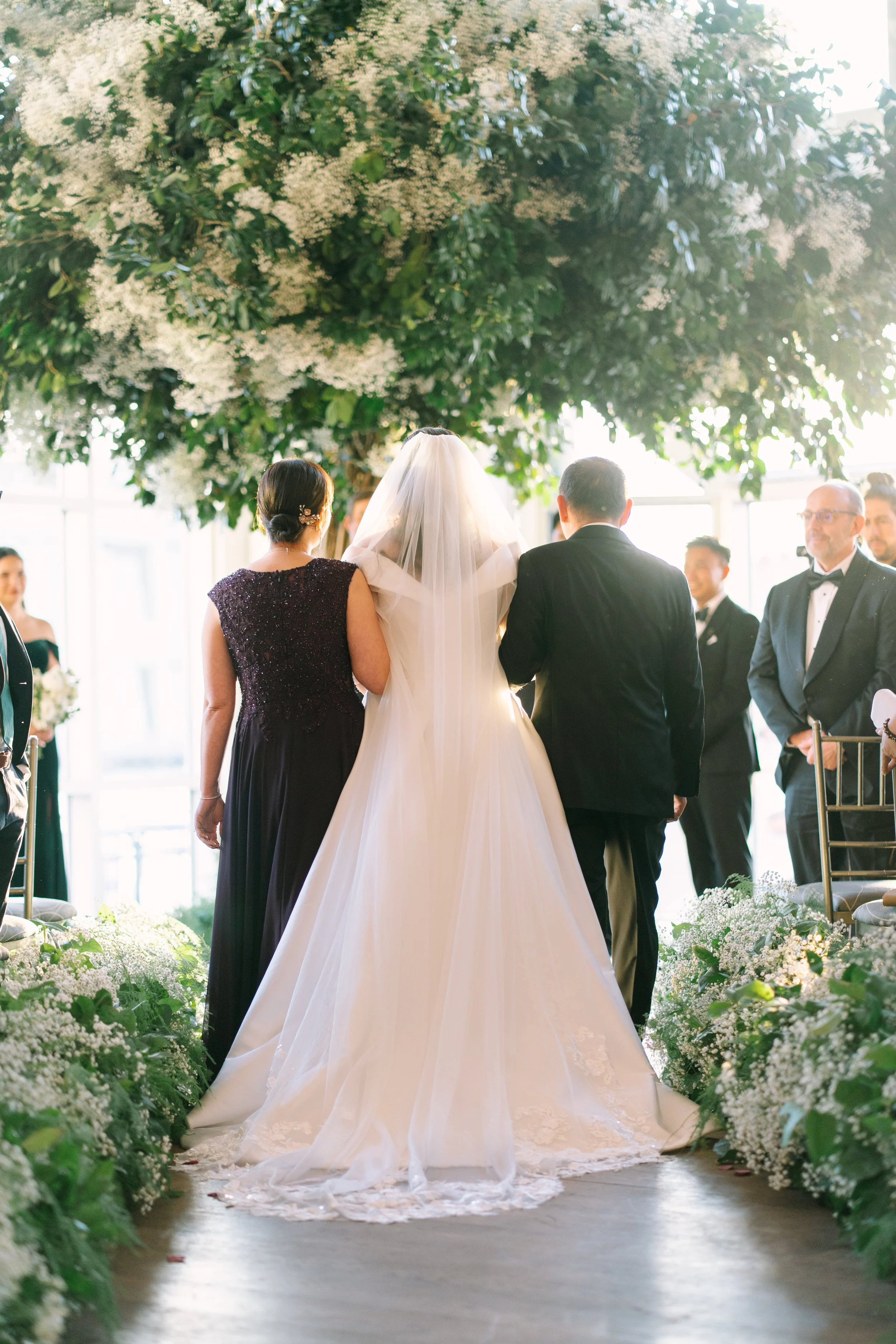 A bride in a white wedding gown walking down the aisle with her parents during a wedding ceremony.