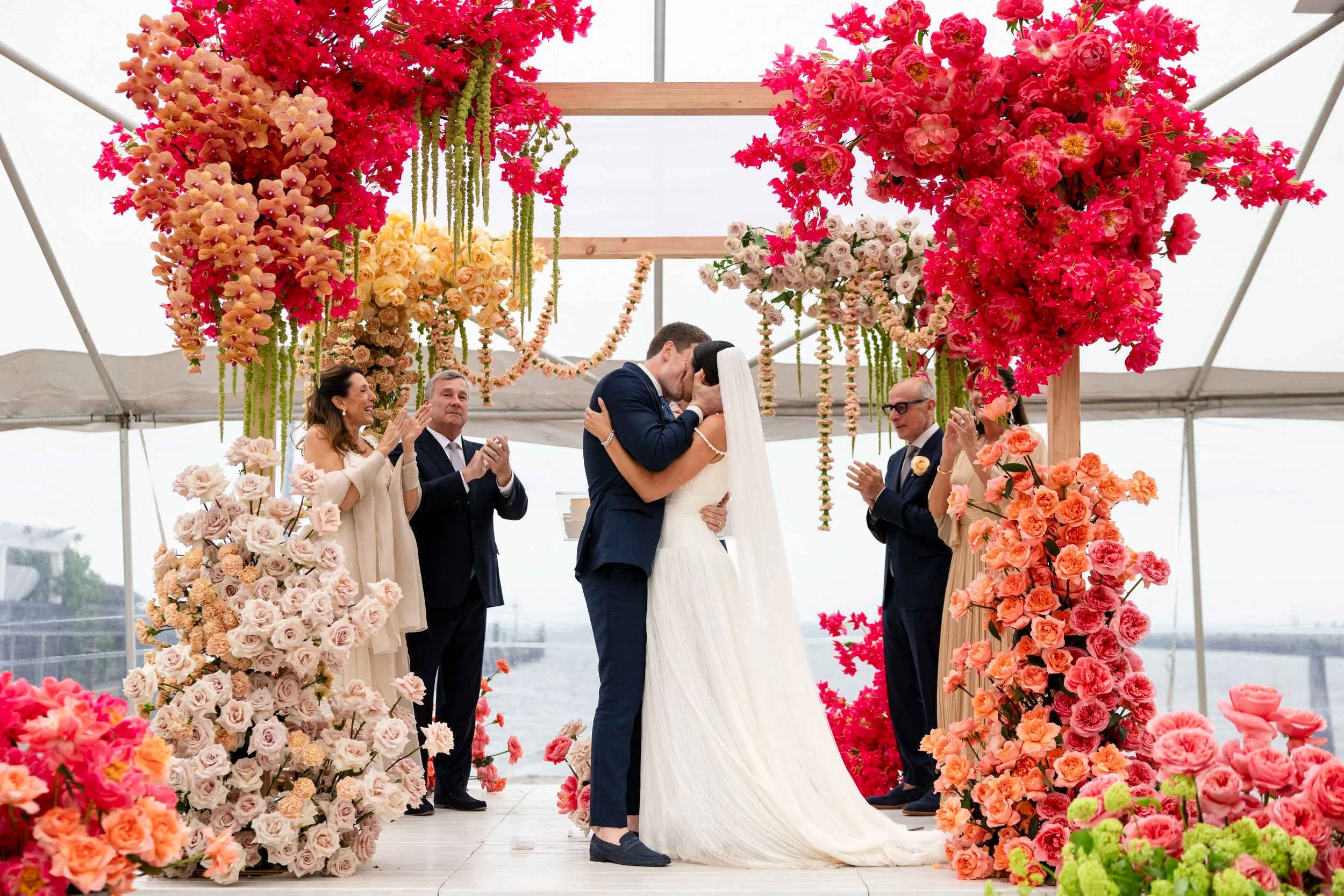 A newlywed couple sharing a kiss during their wedding ceremony, surrounded by colorful pink and peach flowers, with friends and family clapping