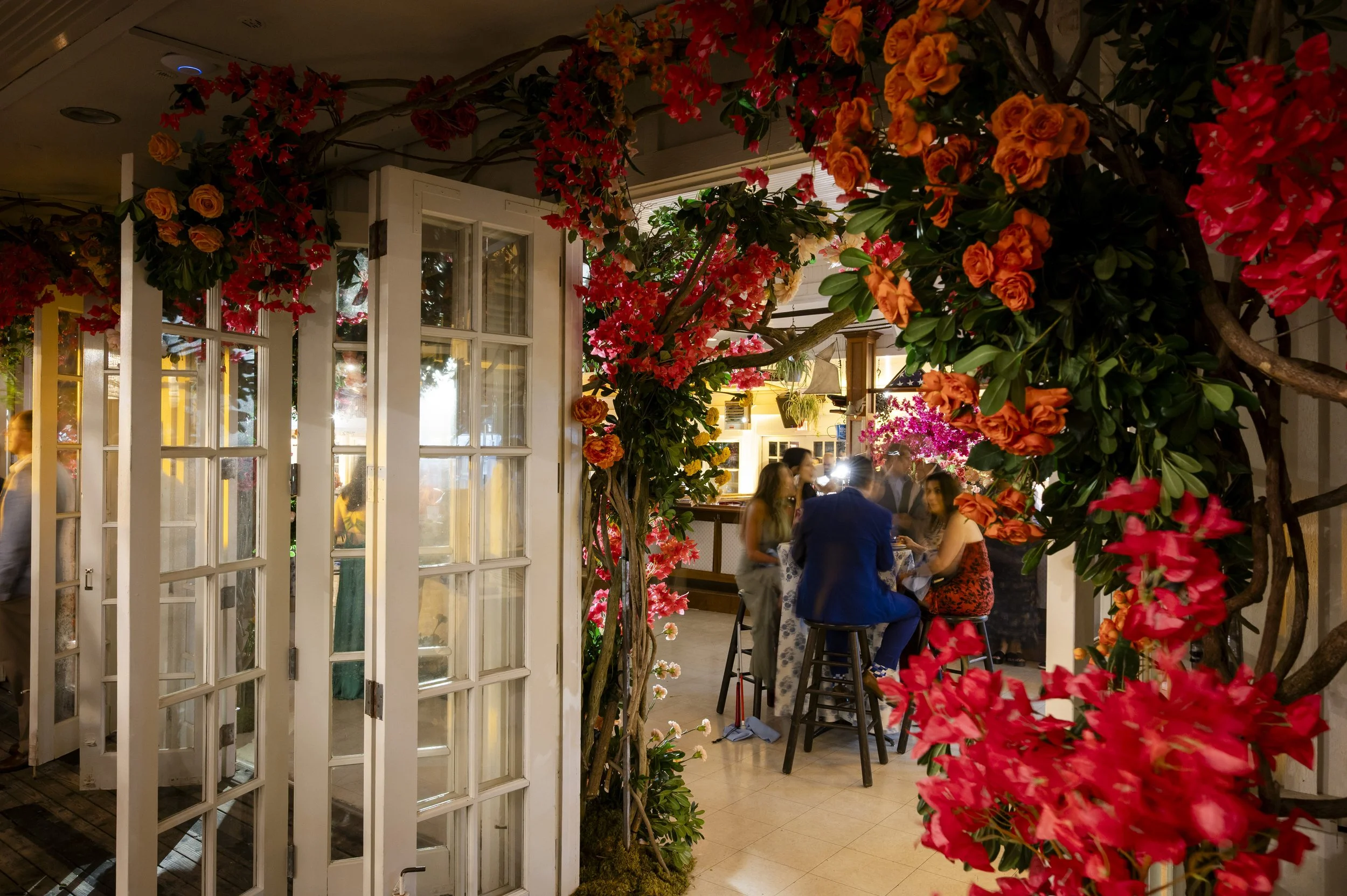 A cozy restaurant or cafe interior decorated with vibrant pink and orange flowers, with patrons sitting at a high table, engaging in conversation.