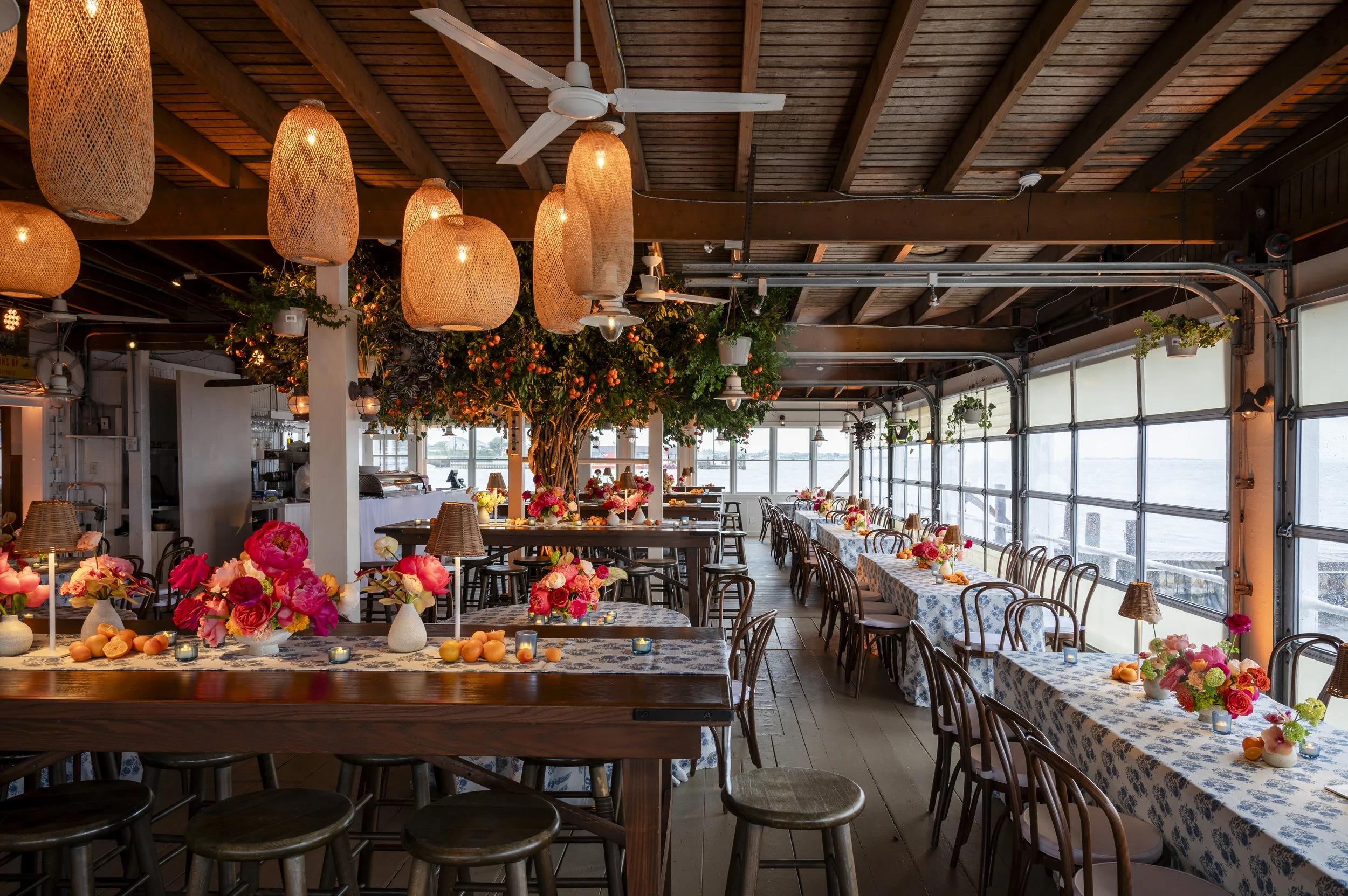 Decorated dining area with wooden ceiling, hanging lights, large window with a view, tables with floral arrangements, candles, and tableware, and a large artificial tree in the center.