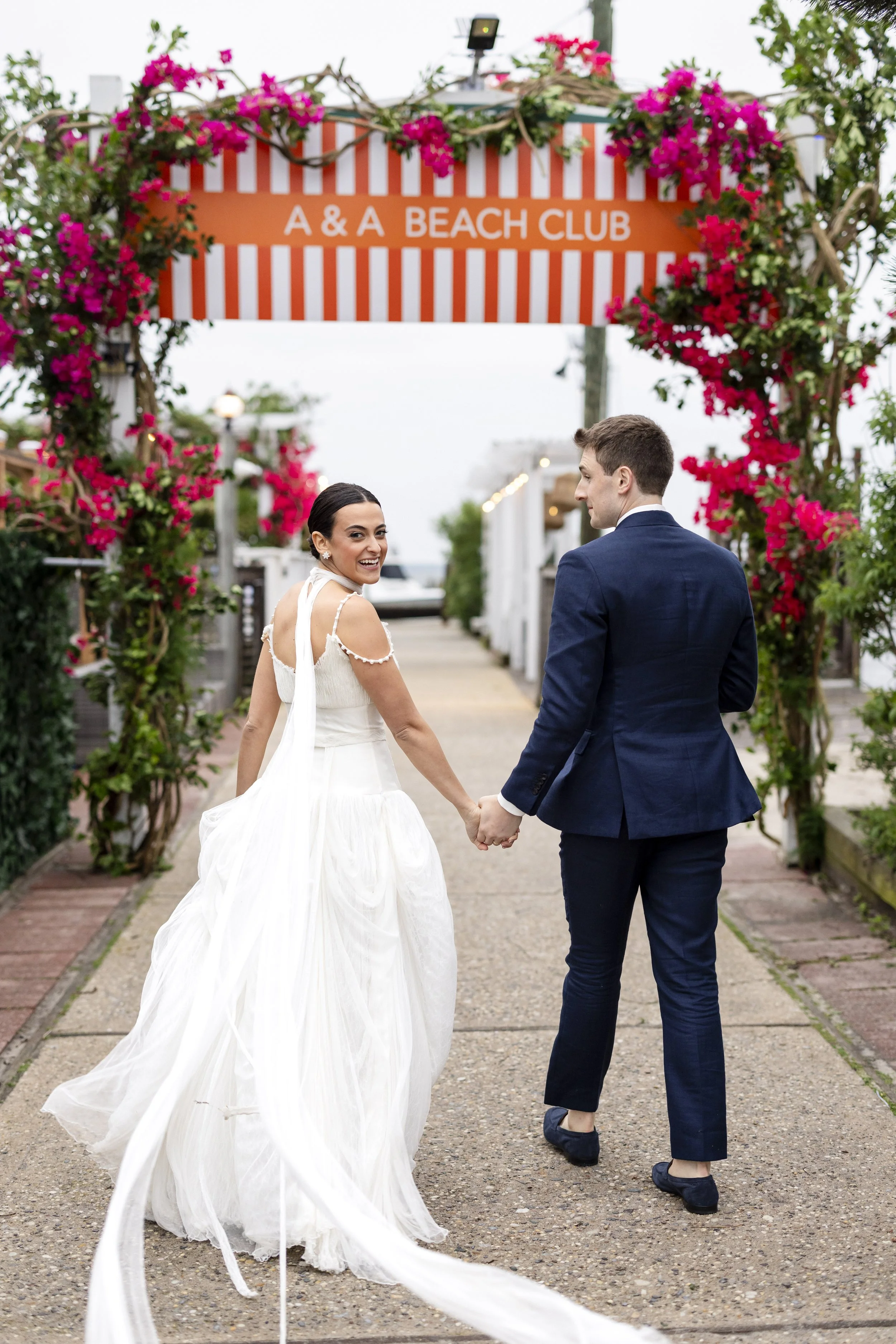 A newlywed couple holding hands and smiling at each other at the entrance of A & A Beach Club, surrounded by pink flowers and greenery.