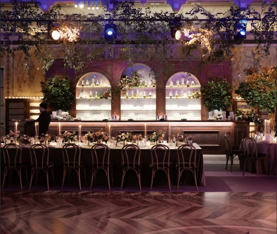 Elegant banquet hall with a long table set for a formal event, floral centerpieces, candles, and a bar with bottles in the background, decorated with greenery and soft lighting.