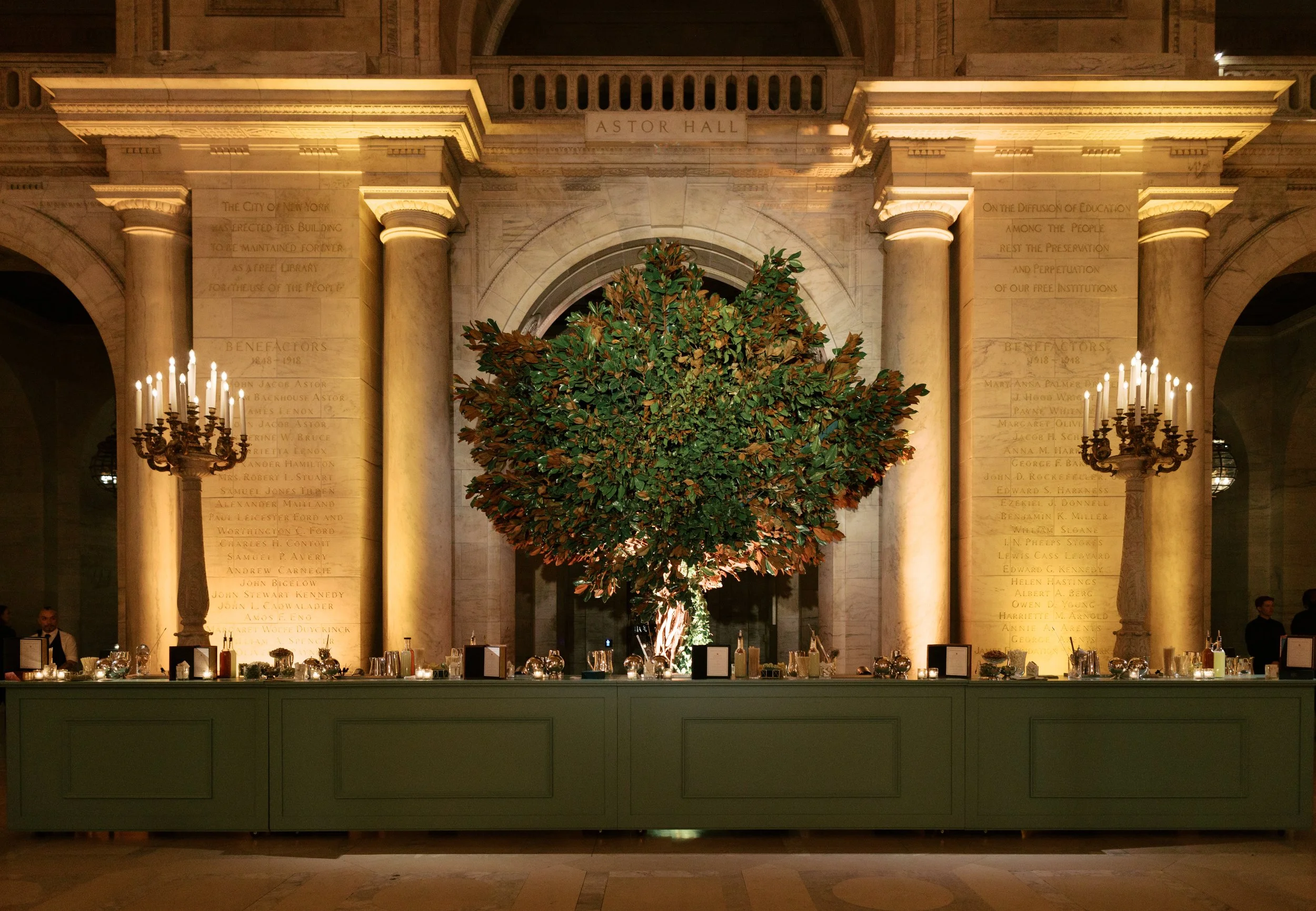 A large indoor space with a prominent green tree in front of a historic wall named Astor Hall, flanked by tall columns with ornate lighting fixtures, and a long table with candles and decor along the front.