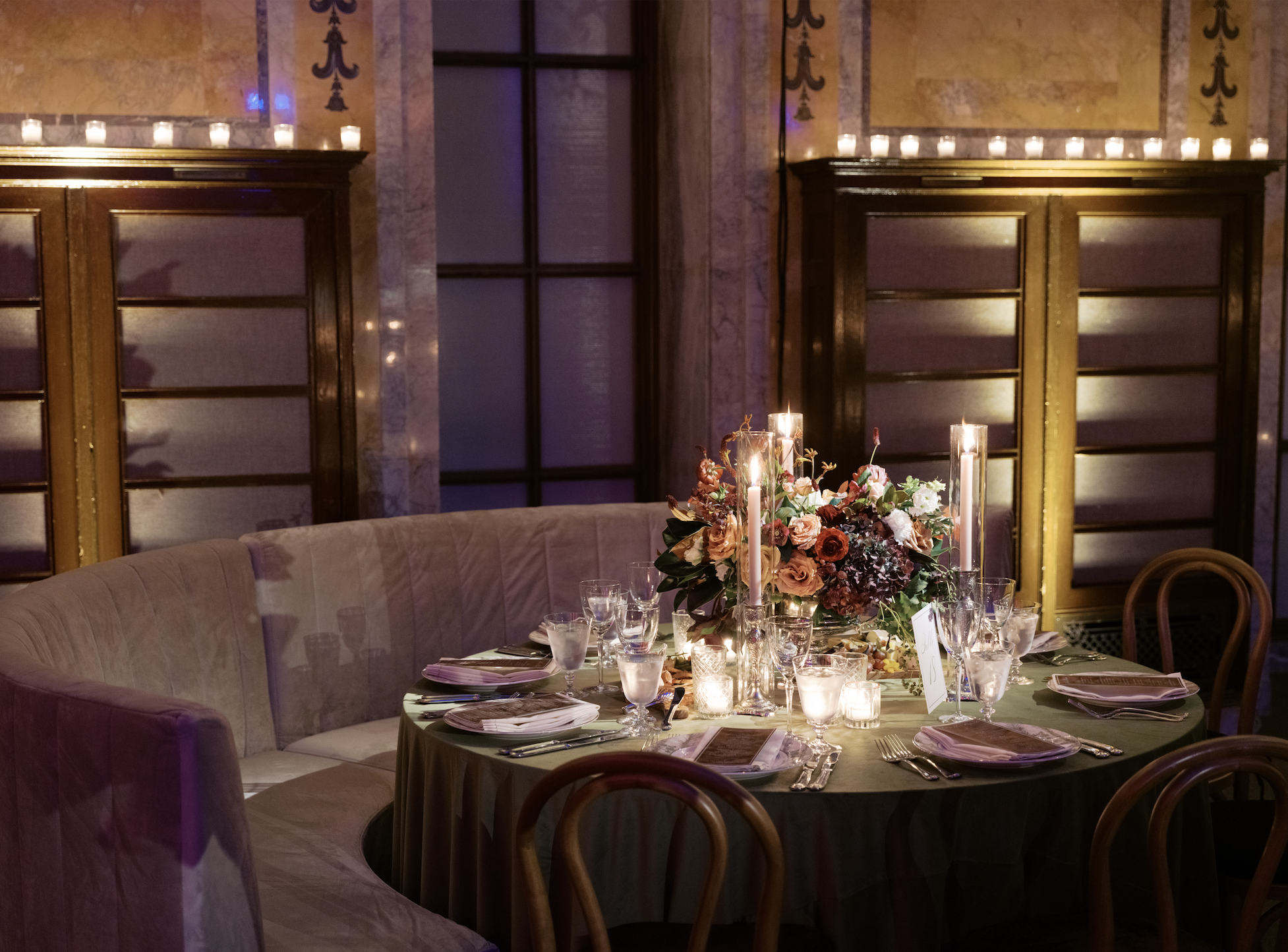 Elegant dining room with a round table set with multiple glasses, plates, silverware, and a large floral centerpiece, surrounded by a curved beige velvet sofa and wooden chairs, featuring candle lighting and decorative wall panels.