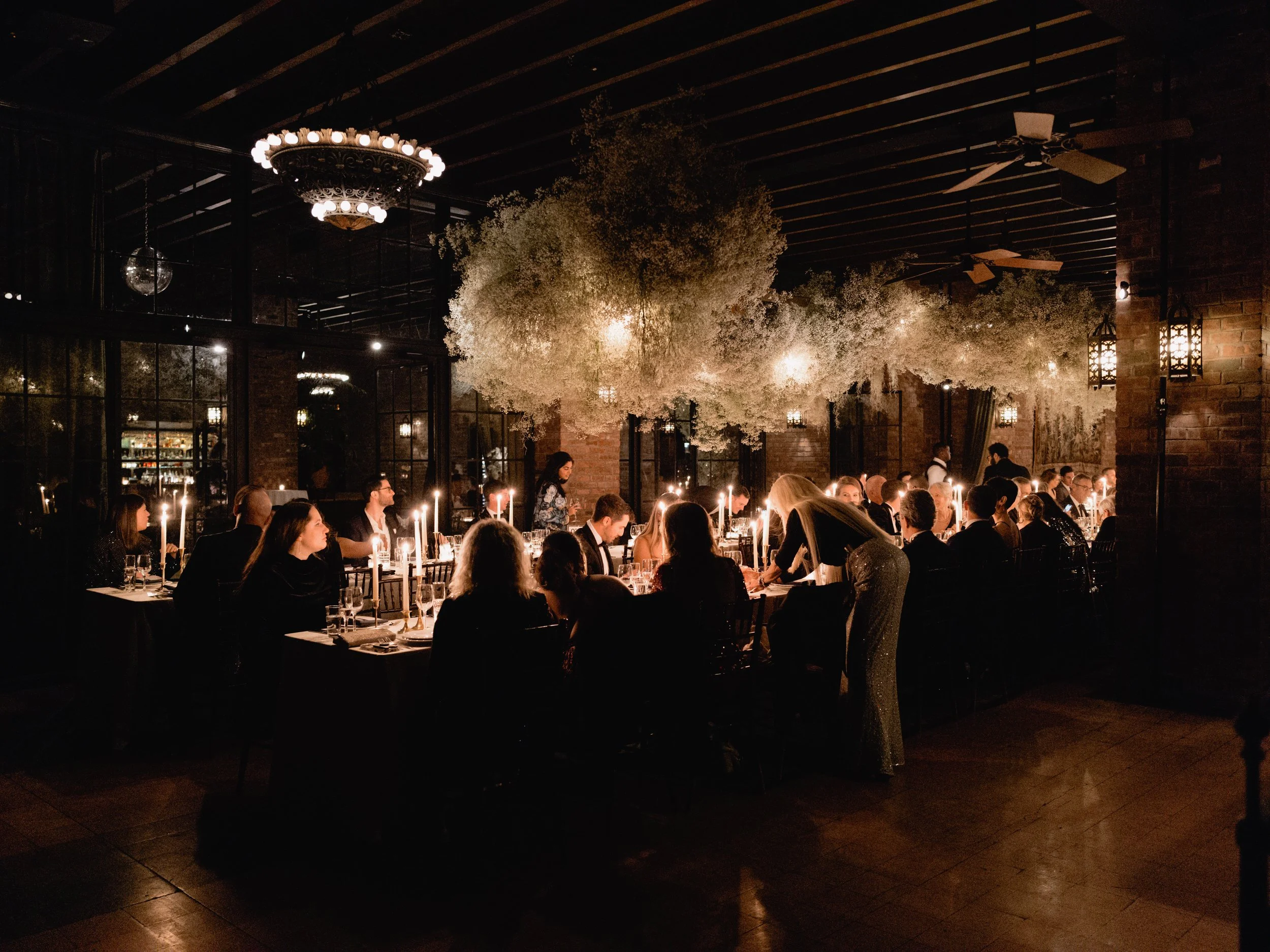 A formal dinner party in a dimly lit restaurant, with guests seated at long tables adorned with candles, surrounded by white floral arrangements and large trees inside the venue. The ceiling has chandeliers and ceiling fans.