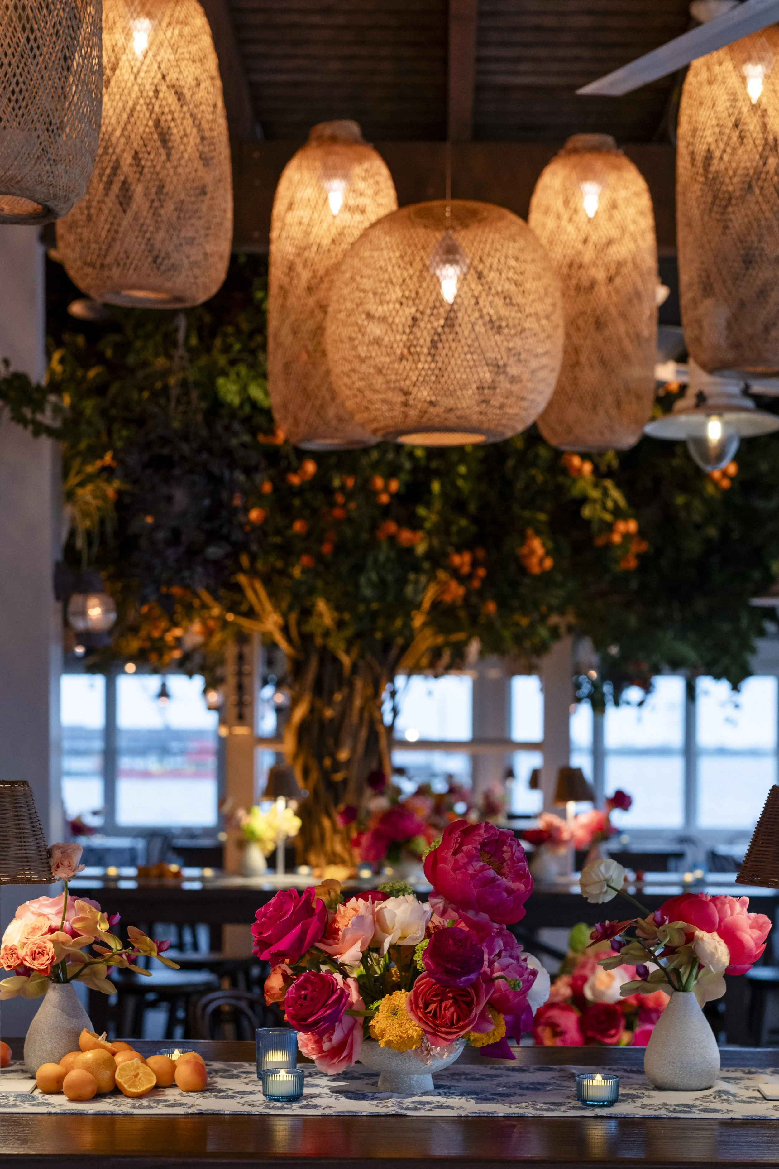 Interior of a restaurant decorated with flower arrangements, hanging woven lamps, and a large tree with orange flowers