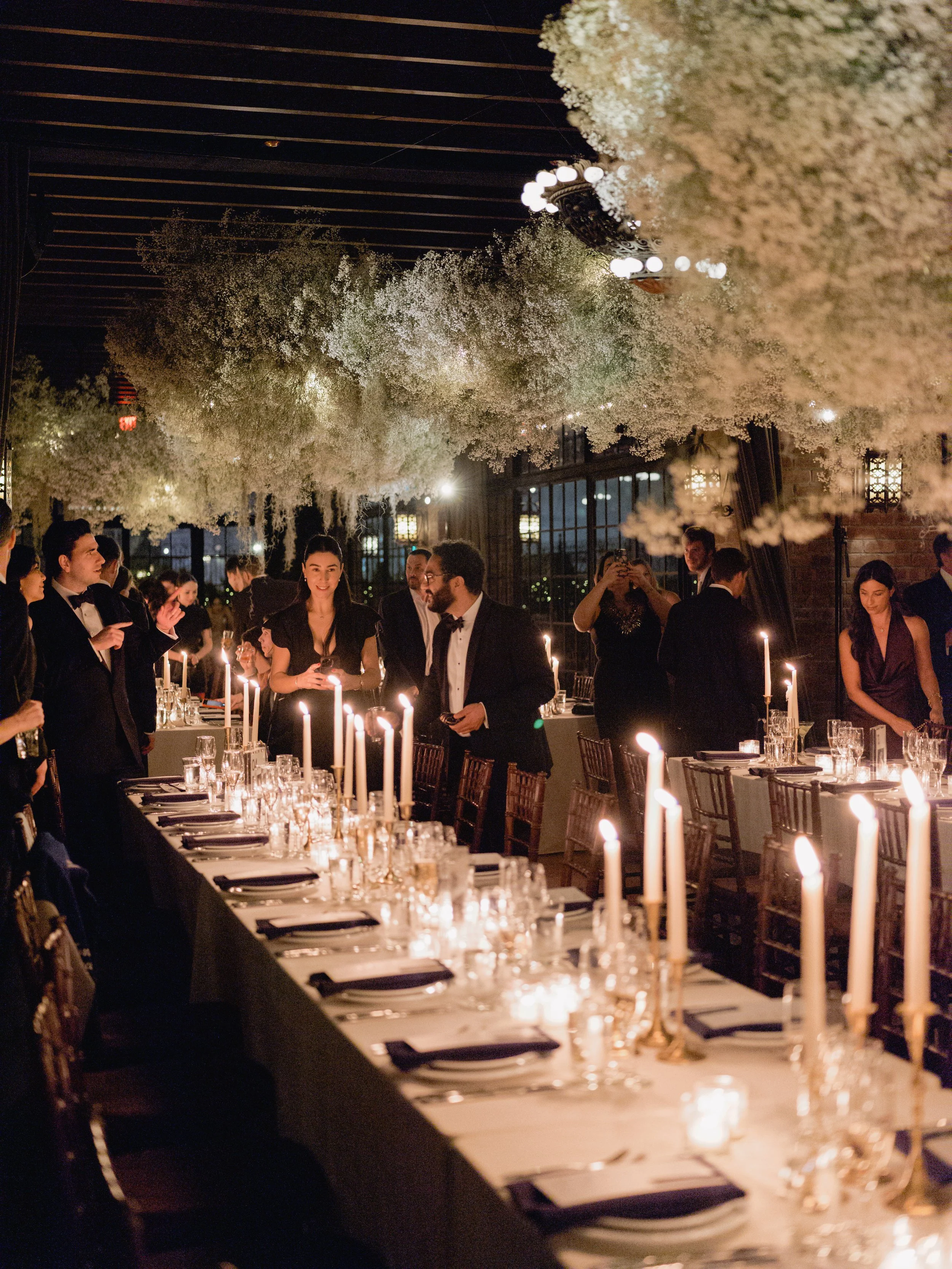 People in formal attire enjoying a dinner party with a long table, candlelit decor, and hanging floral arrangements in an elegant venue.
