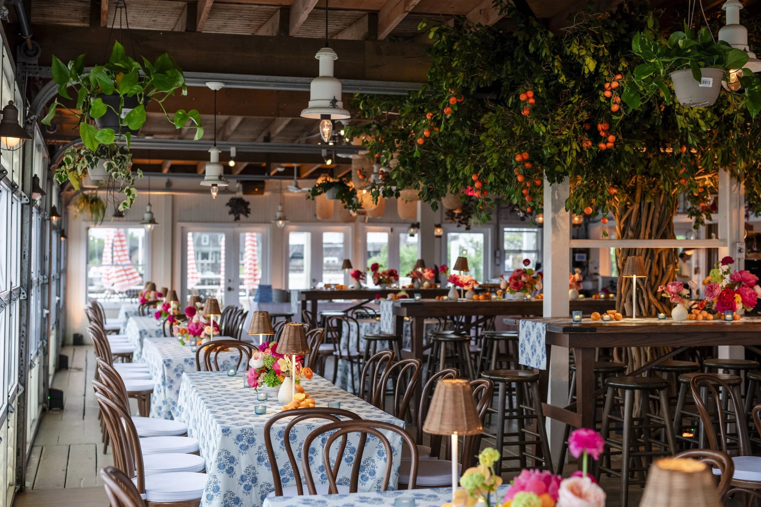 Restaurant interior decorated with pink and orange flowers on tables, orange fruit hanging from a large tree, and string lights hanging from the ceiling.