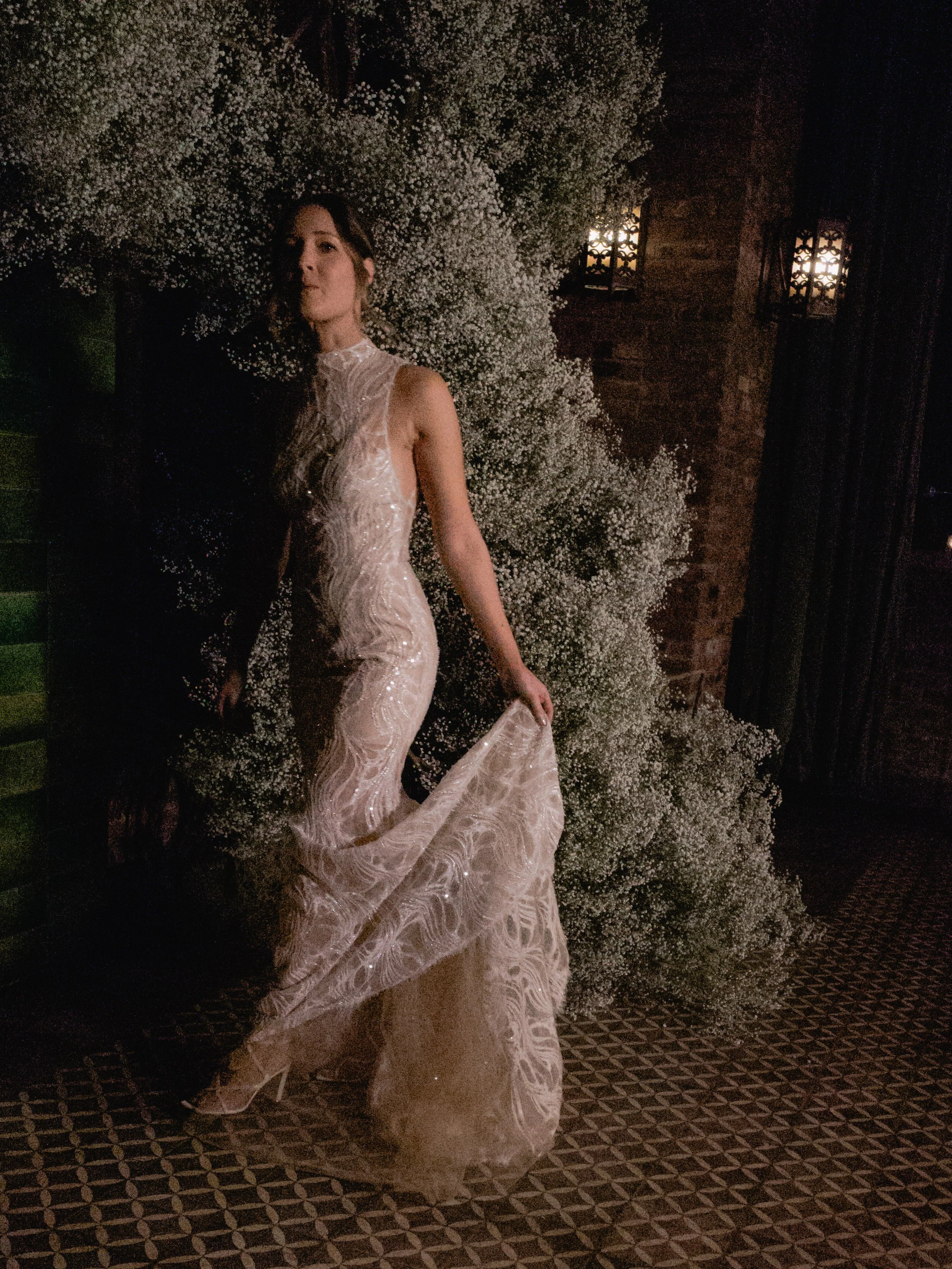 A woman in an elegant, light-colored, patterned gown poses in front of a large white floral arrangement, dimly lit by wall sconces, with a brick wall and patterned floor visible.