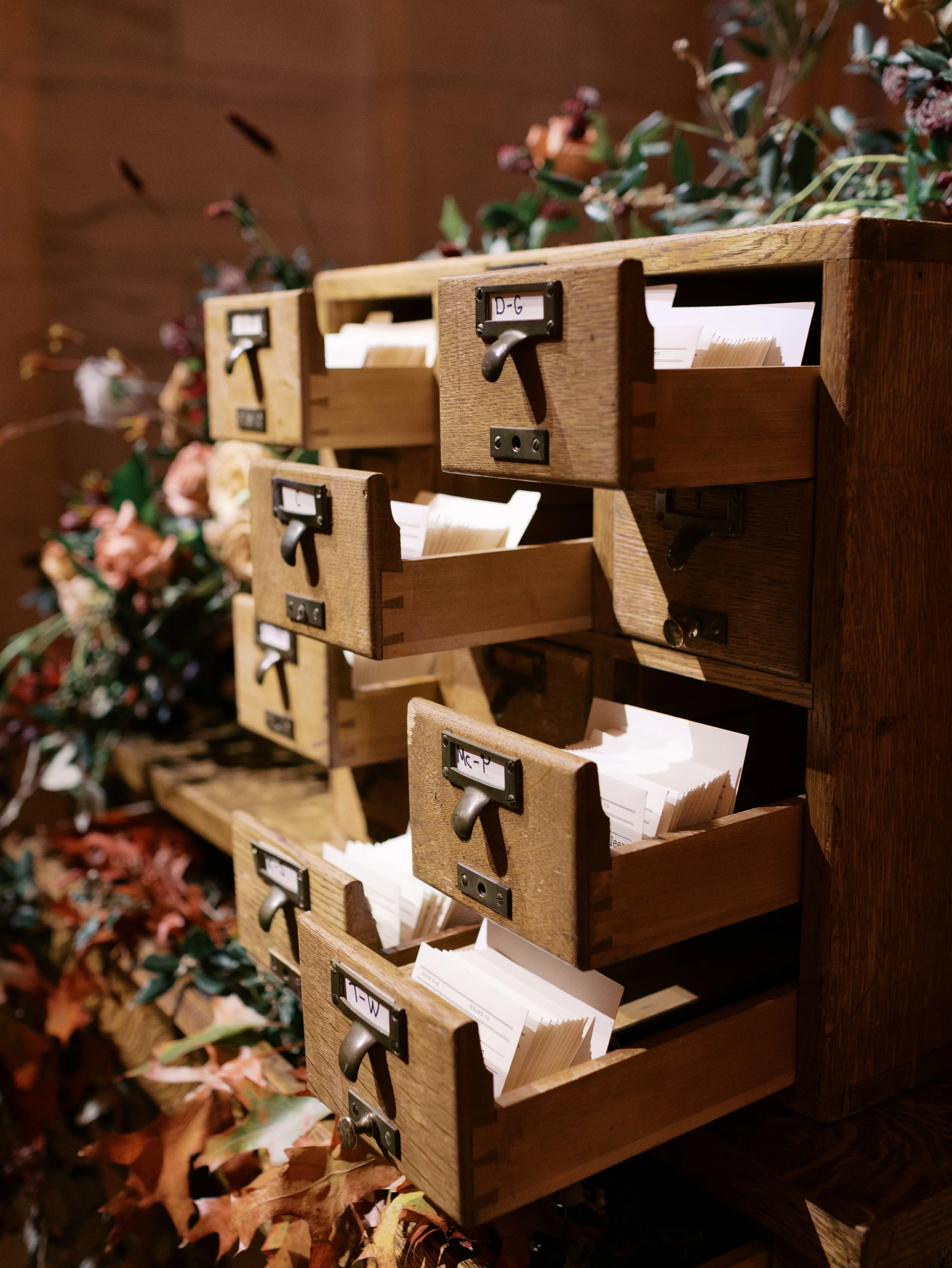 Wooden mail organizer with labeled drawers containing stacks of envelopes, placed on a table with autumn leaves and floral decorations in the background.