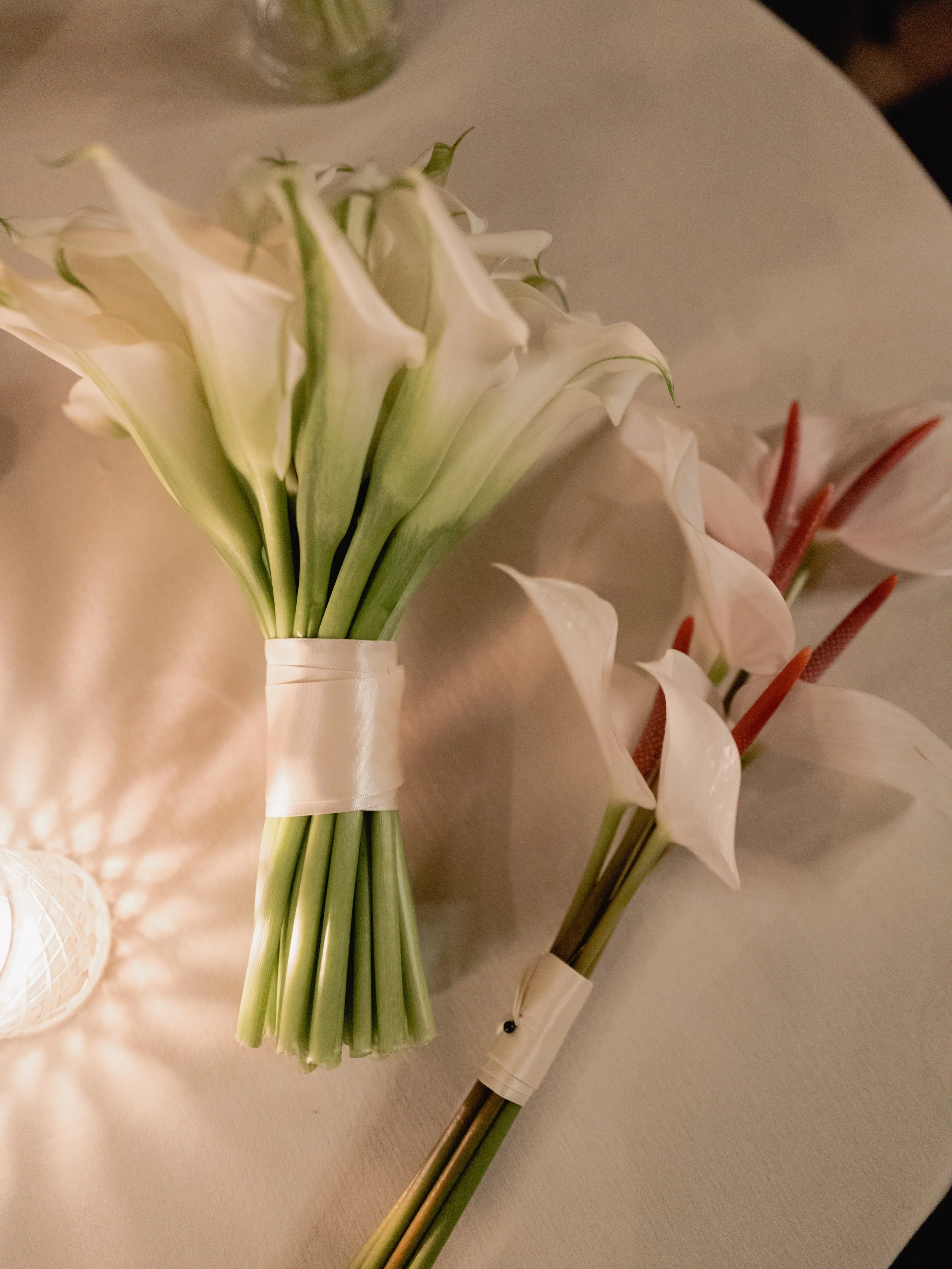Bouquet of white calla lilies wrapped with white ribbon, lying on a light-colored surface.