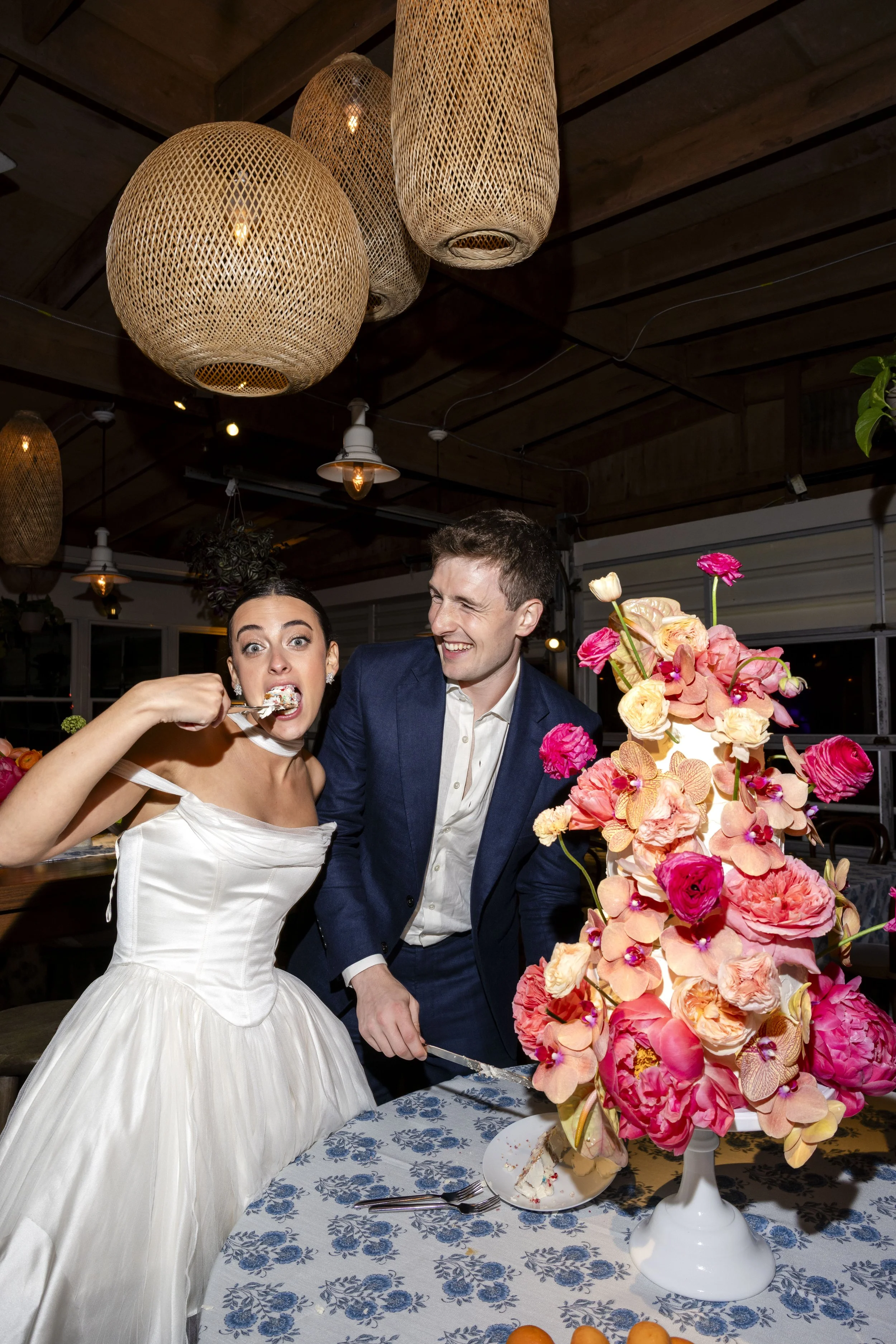 A woman in a white dress feeds cake to a man in a dark blue suit during a celebration, with a large pink floral centerpiece on a blue and white patterned tablecloth, under warm hanging light fixtures in a rustic interior.