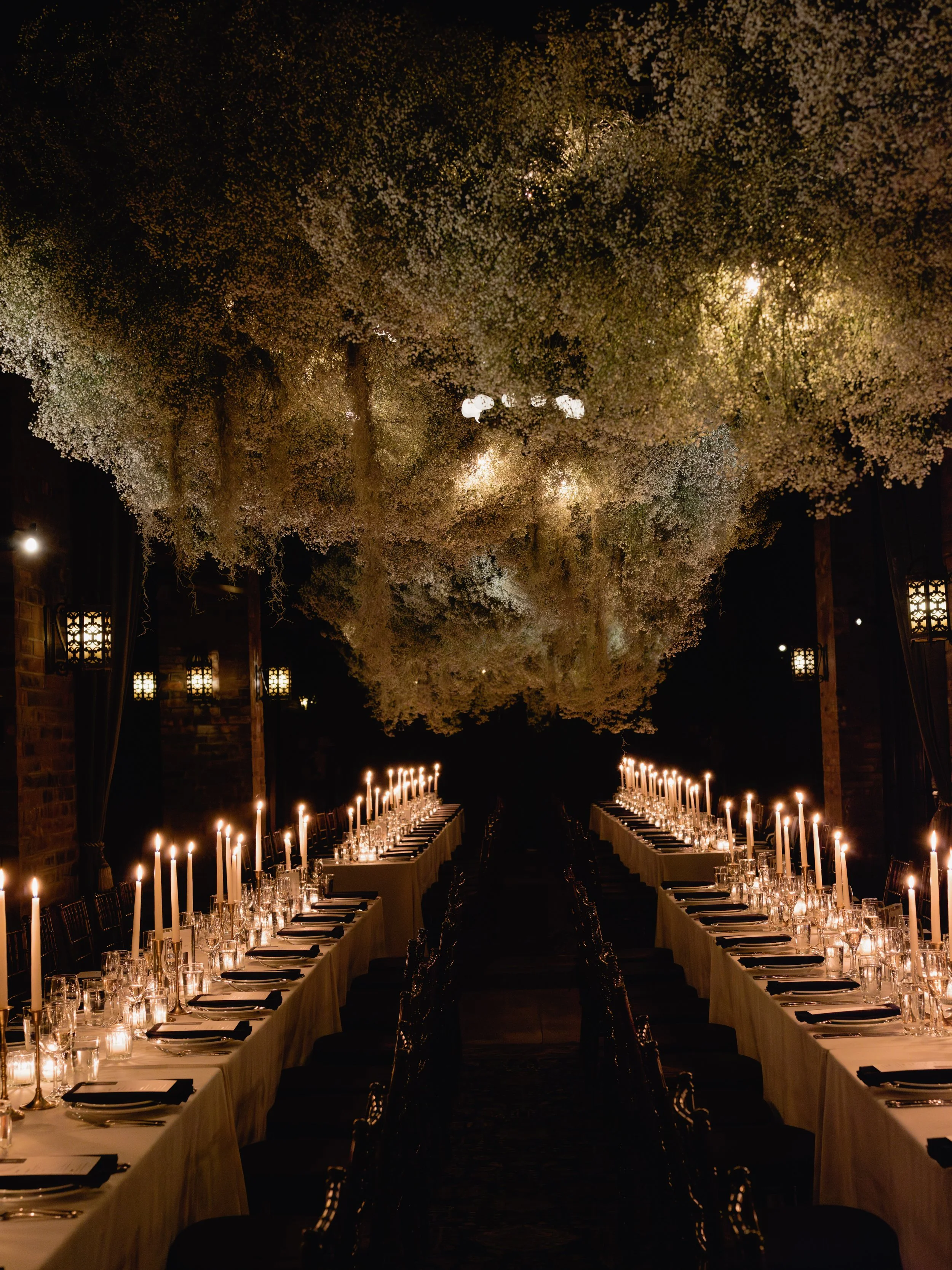 Elegant dining room set for a formal event with long tables, lit candles, and a decorative ceiling with hanging flowers or foliage.