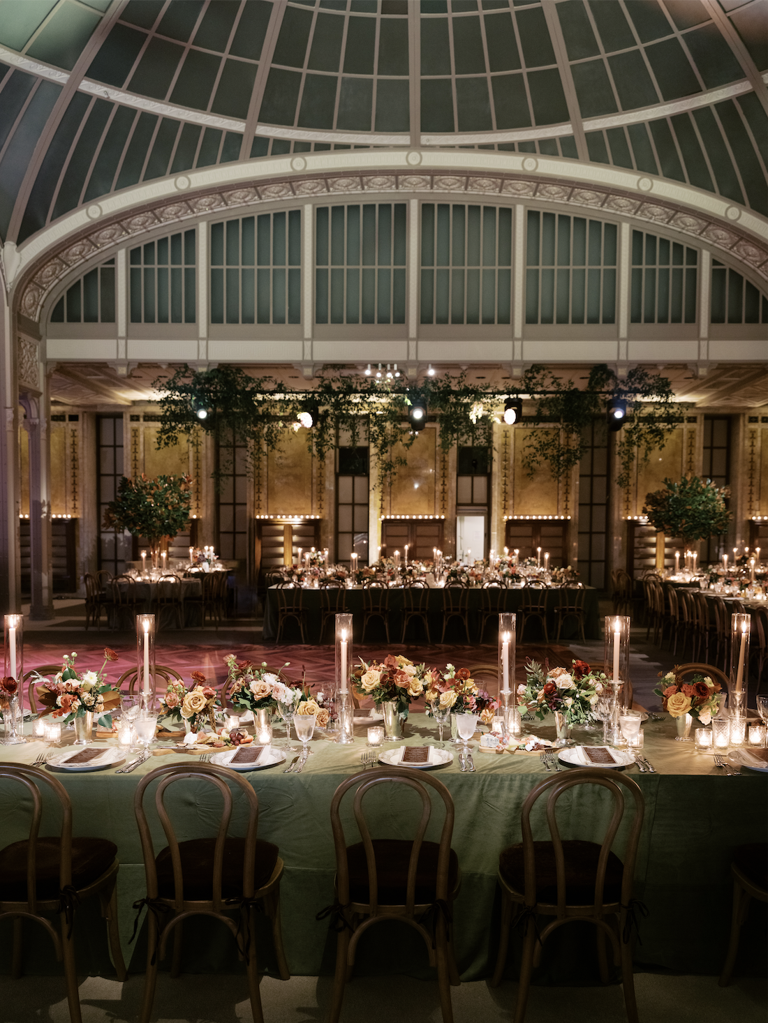 Elegant banquet hall with round and rectangular tables decorated with floral centerpieces, candles, and table settings, under a glass-domed ceiling with hanging greenery and warm lighting.